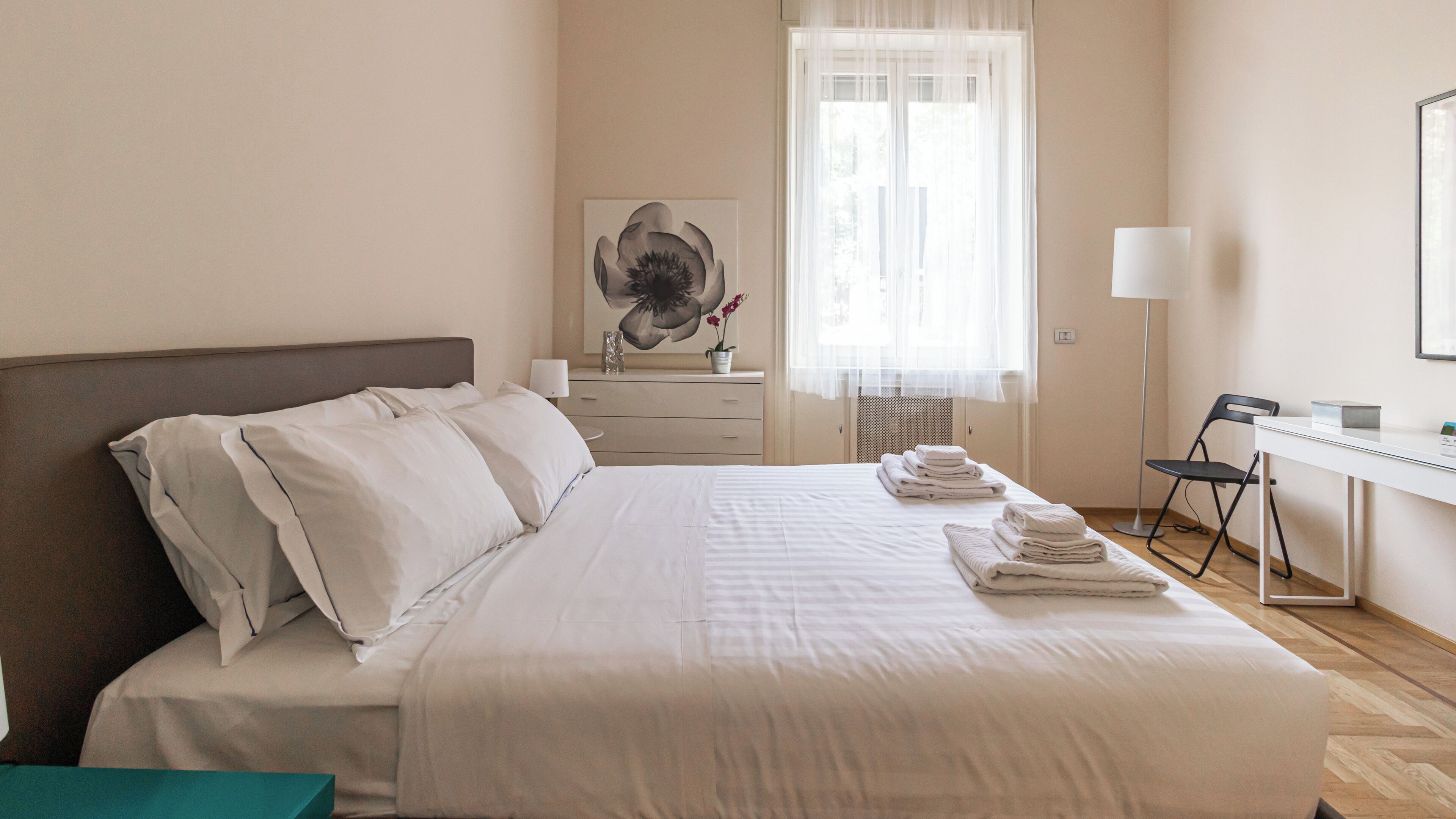 Una cama bien tendida con sábanas blancas, una mesita de noche con lámpara, un escritorio con silla y una ventana con cortinas translúcidas.