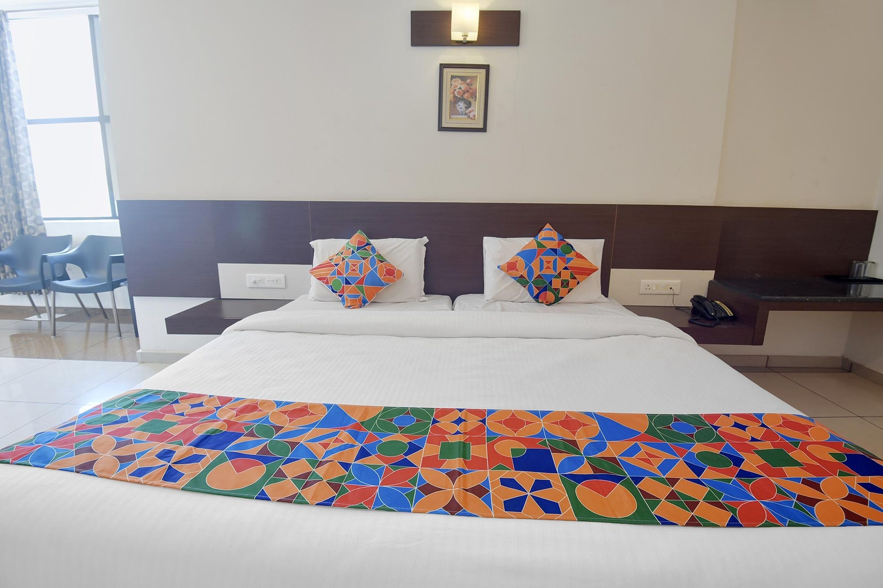 Habitación de hotel con una cama grande, dos almohadas con diseños geométricos, una mesita de noche, una silla y un ventanal con cortinas.
