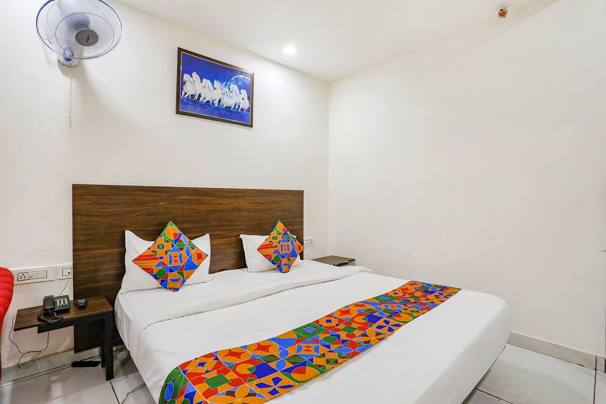 Een hotelkamer met een houten hoofdeinde, een bed met wit beddengoed, kleurrijke kussens, een nachtkastje met telefoon en oplader, een aan de muur hangend schilderij en een vloerventilator.