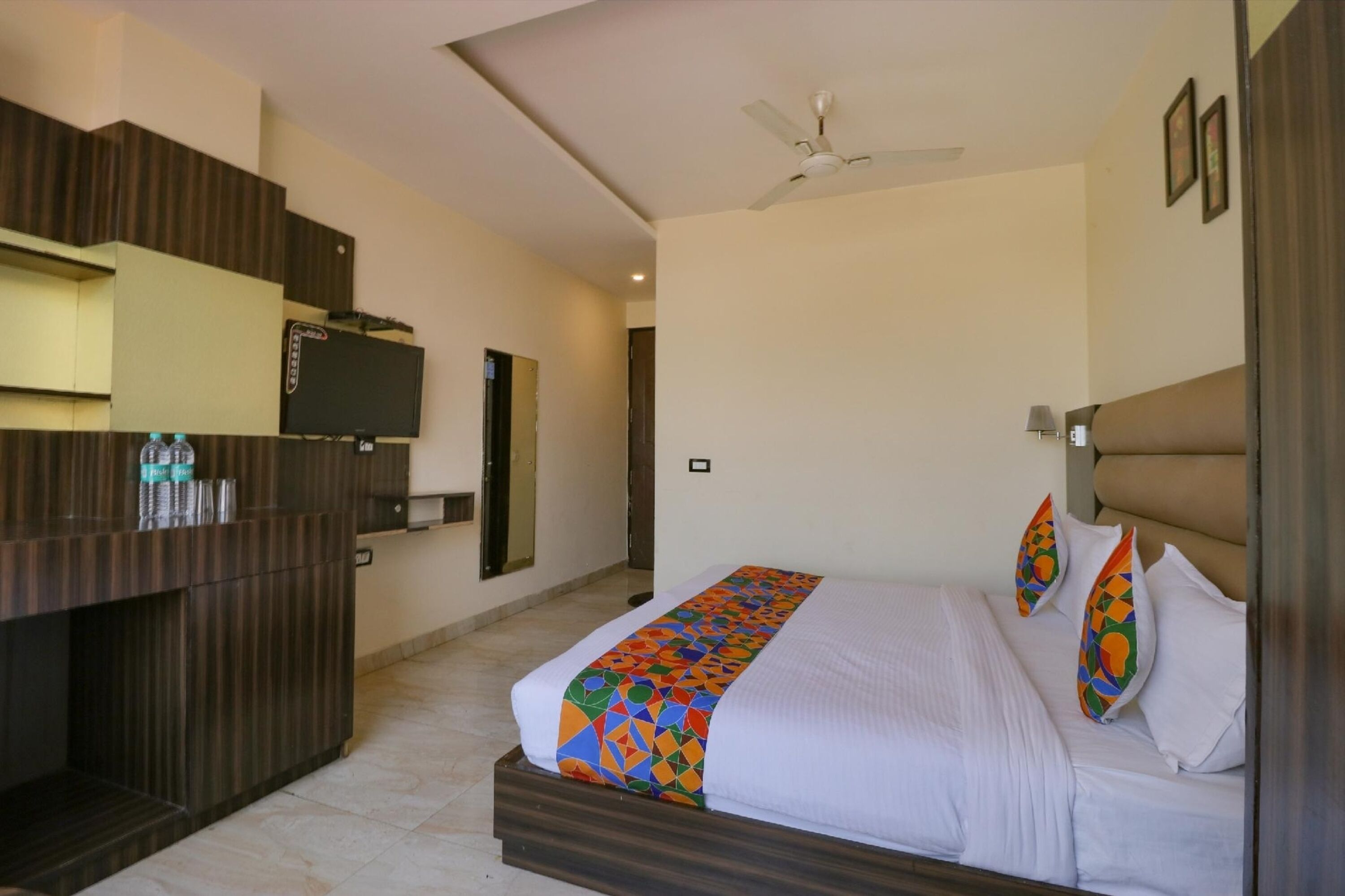 A hotel room with a bed, a TV, a ceiling fan, and a minibar.