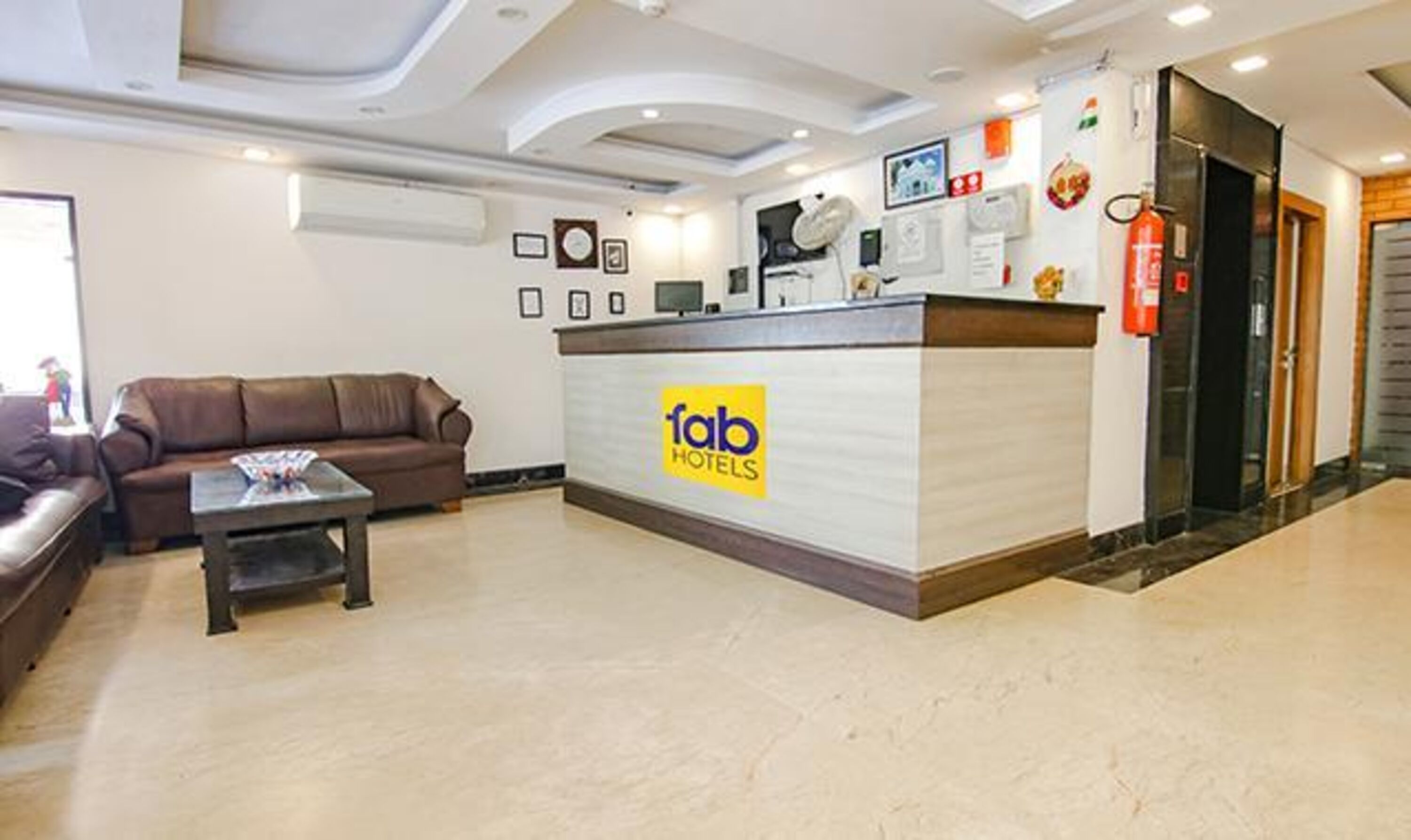 A lobby with a reception desk labeled 'fab hotels', a brown leather sofa, a coffee table, and a fire extinguisher.