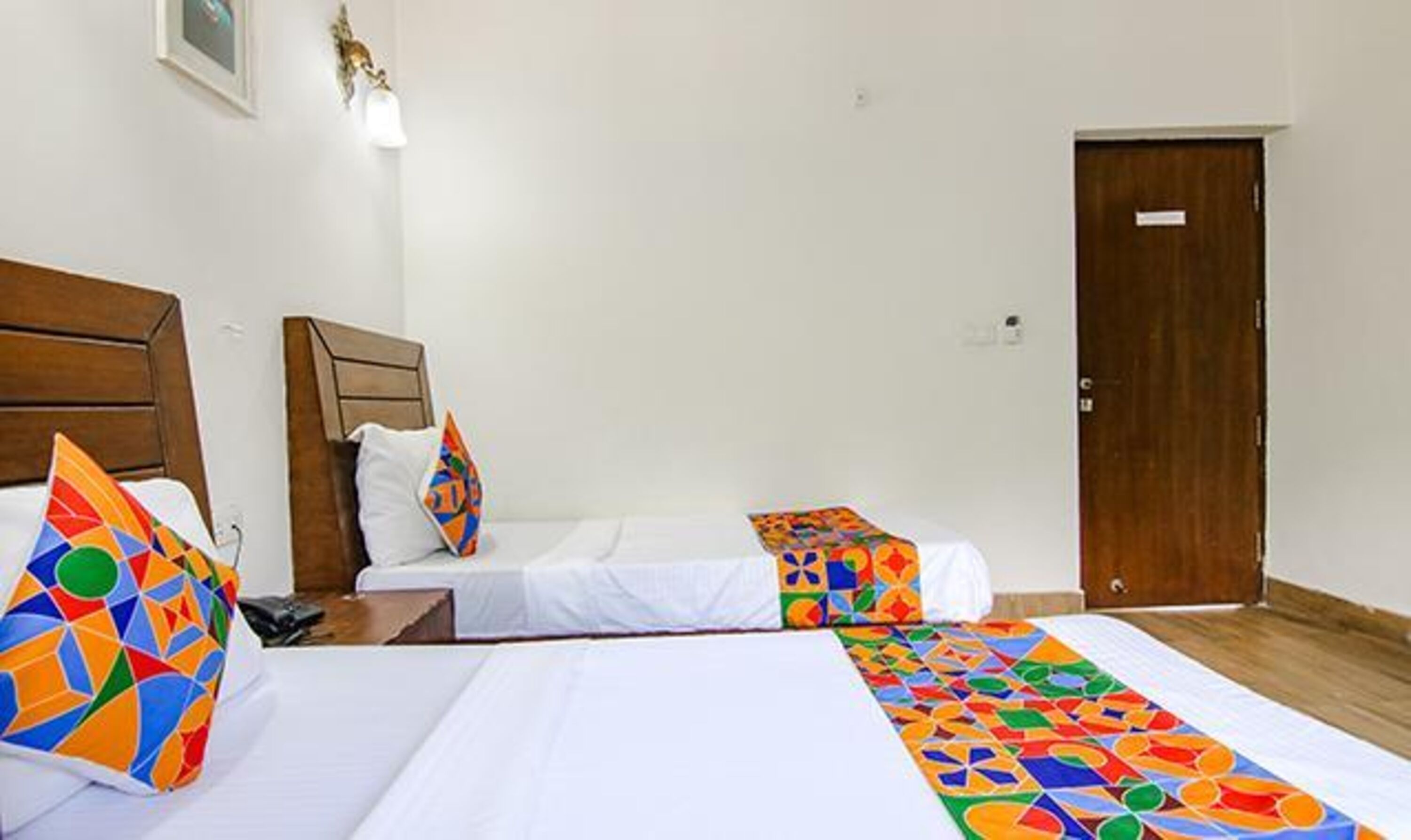 A hotel room with two single beds, a wooden headboard, a white wall, a door, and a small table with a lamp.