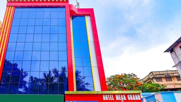 A multi-story building with a prominent glass facade, a vertical red and yellow stripe, and a sign for "HOTEL MEGA GRAND". - FabHotel The Crown Comforts (Bidhannagar)