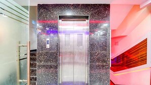 An elevator with a metallic finish and a control panel, surrounded by a speckled wall and a staircase with a red and orange gradient. - FabHotel The Crown Comforts (Bidhannagar)