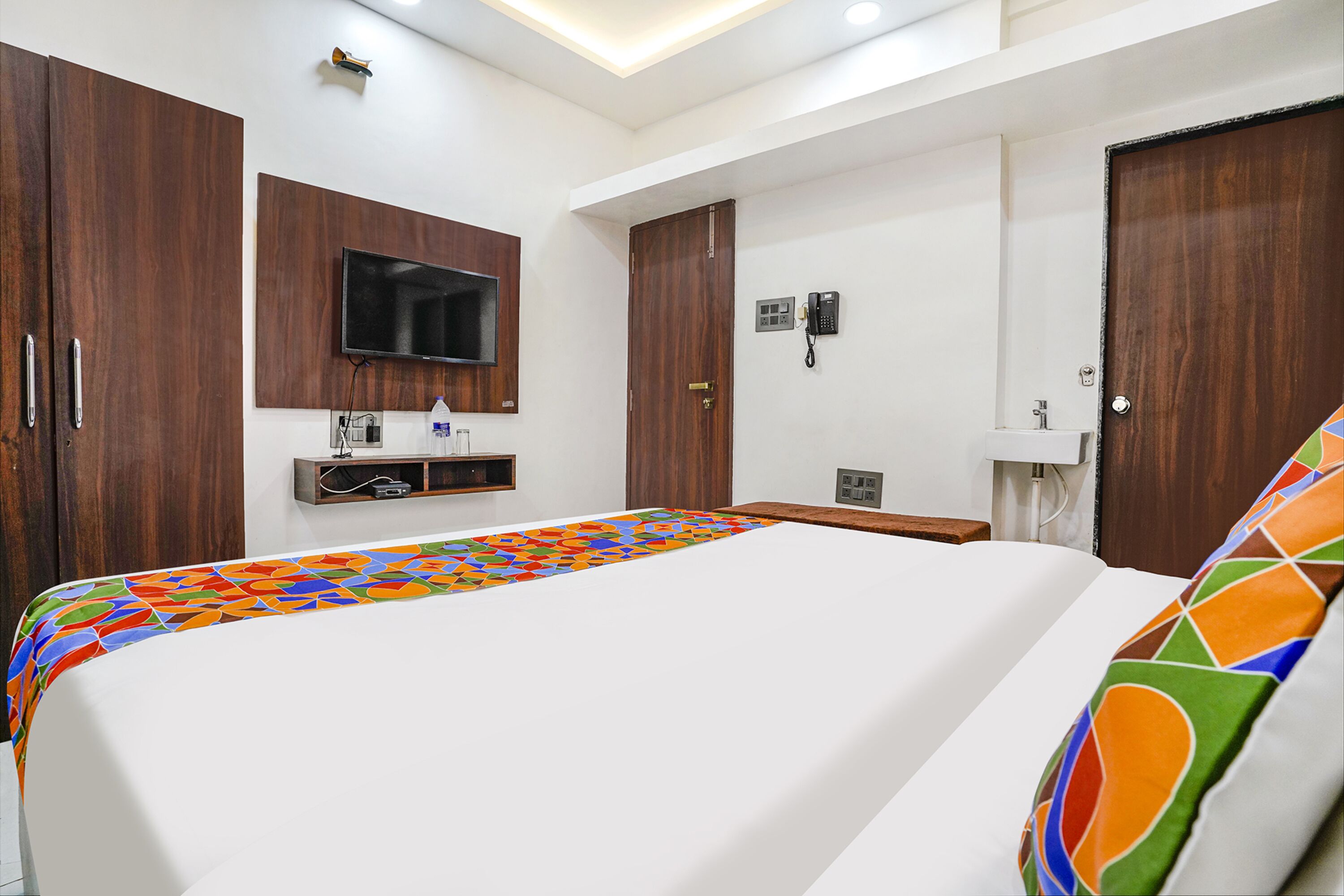 A hotel room with a large bed, wooden wardrobe, a mounted TV, a telephone, and a small desk with a chair.