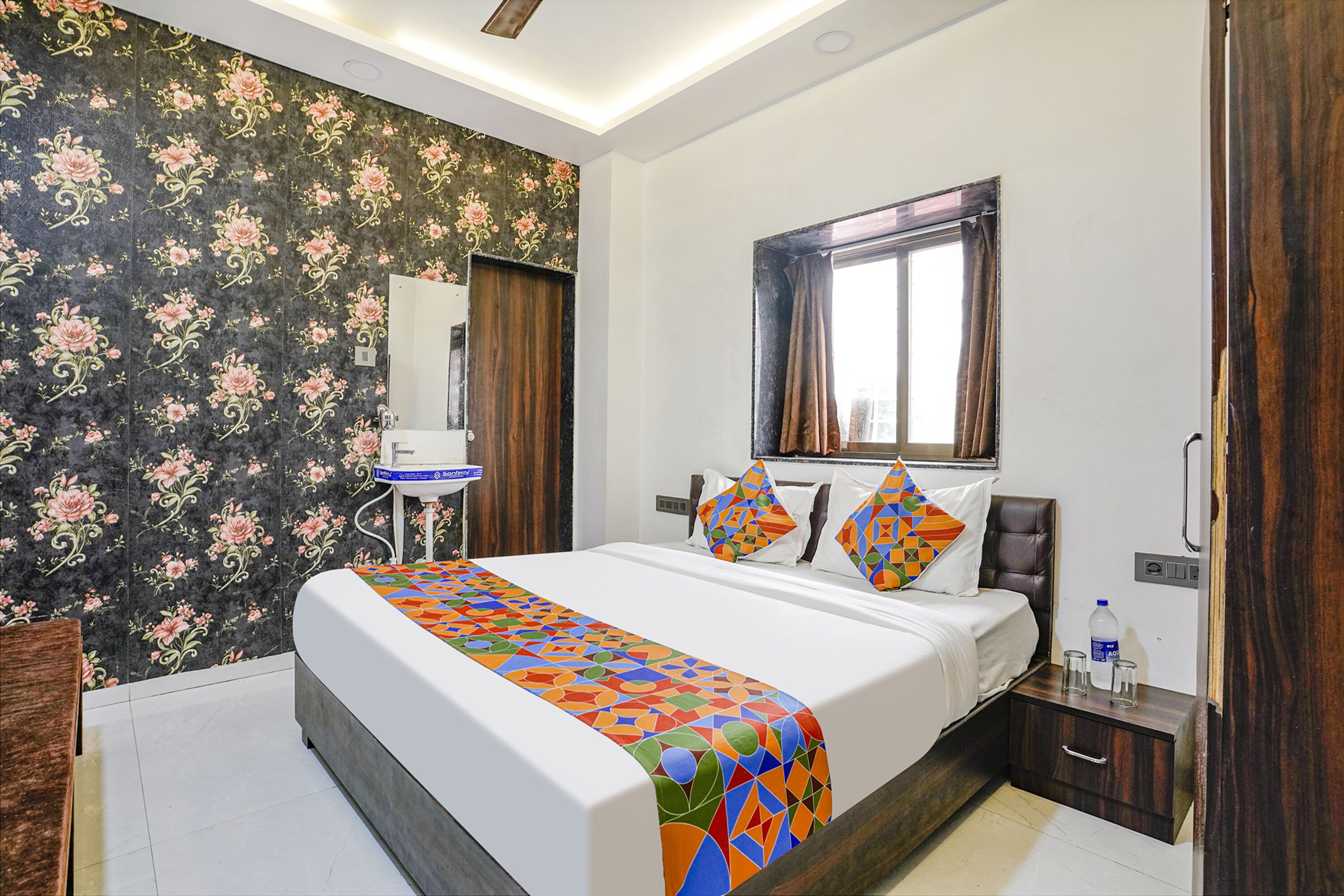 A hotel room with a bed, a bedside table, a mirror, and a window with curtains.