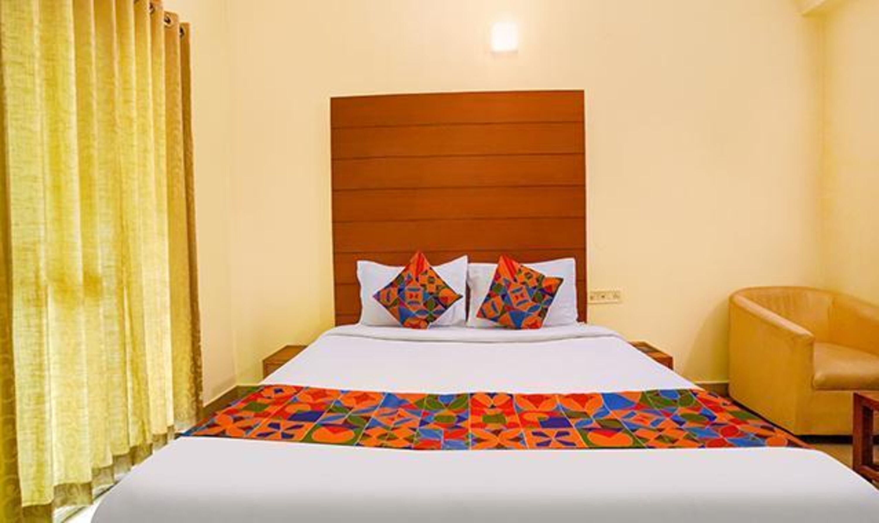 A hotel room with a wooden headboard, a bed with a colorful geometric-patterned bedspread, two decorative pillows, a yellow armchair, and a curtain.