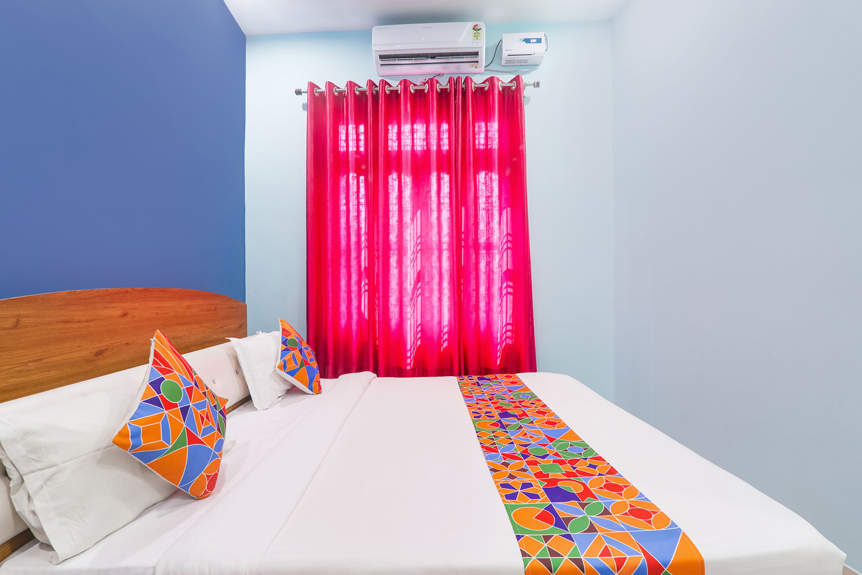 Ein Hotelzimmer mit einem Bett, einem bunten Kissen, einer weißen Bettdecke, einem roten Vorhang und einer Klimaanlage an der Wand.