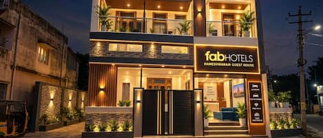 A modern two-story guest house with a well-lit entrance, a signboard, and a gated driveway.