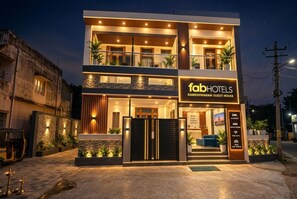 A modern two-story guest house with a well-lit entrance, a signboard, and a gated driveway.