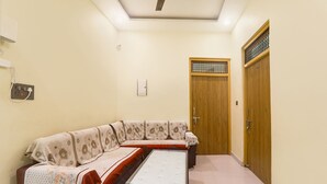 A living room with a sofa, a glass-top coffee table, a wooden door, and a wall-mounted light fixture. - FabHotel Rameshwaram Guest House - 500m From Ram Mandir (Faizabad)