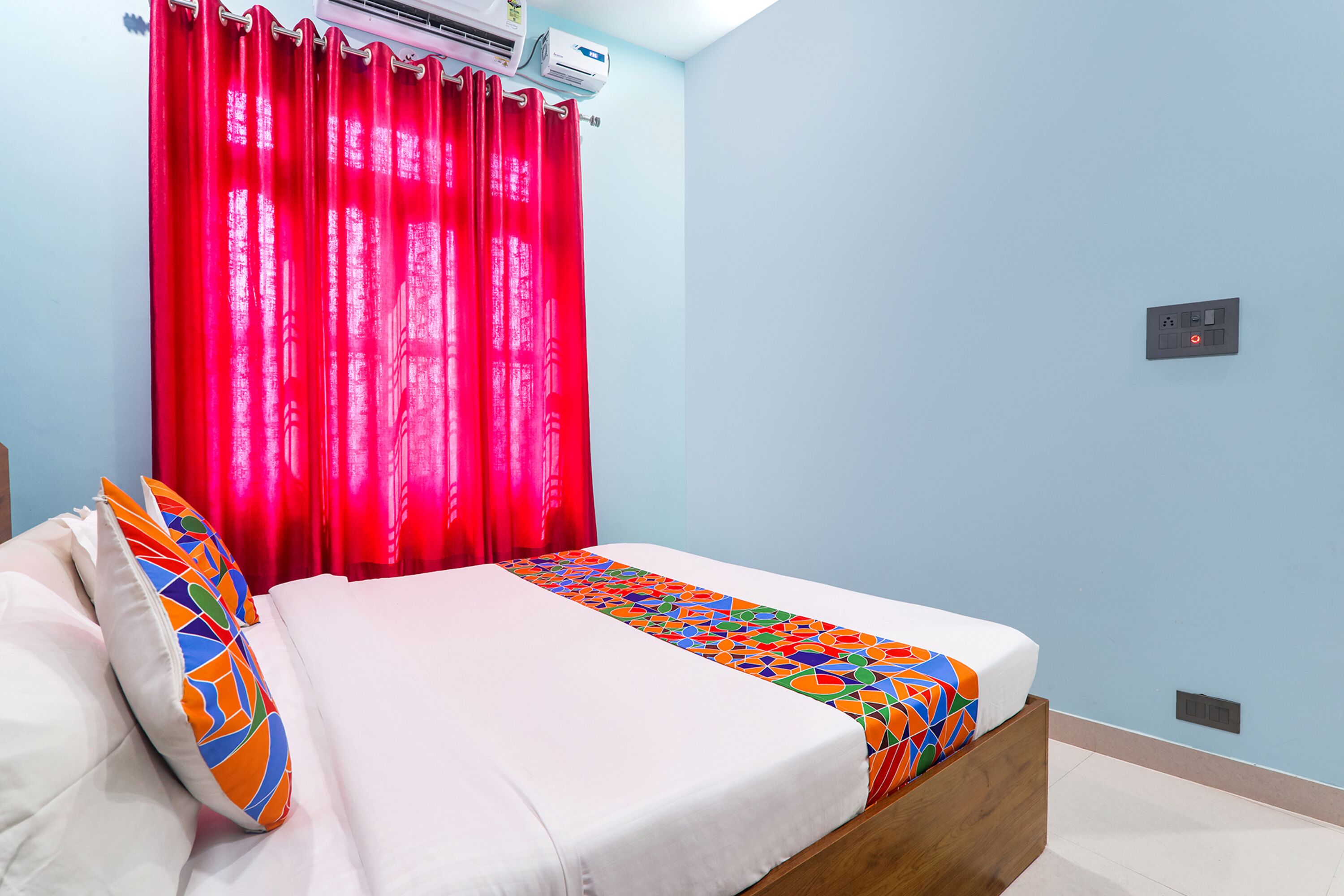 Un dormitorio con una cama, un edredón colorido, una cortina roja y un aire acondicionado instalado en la pared.