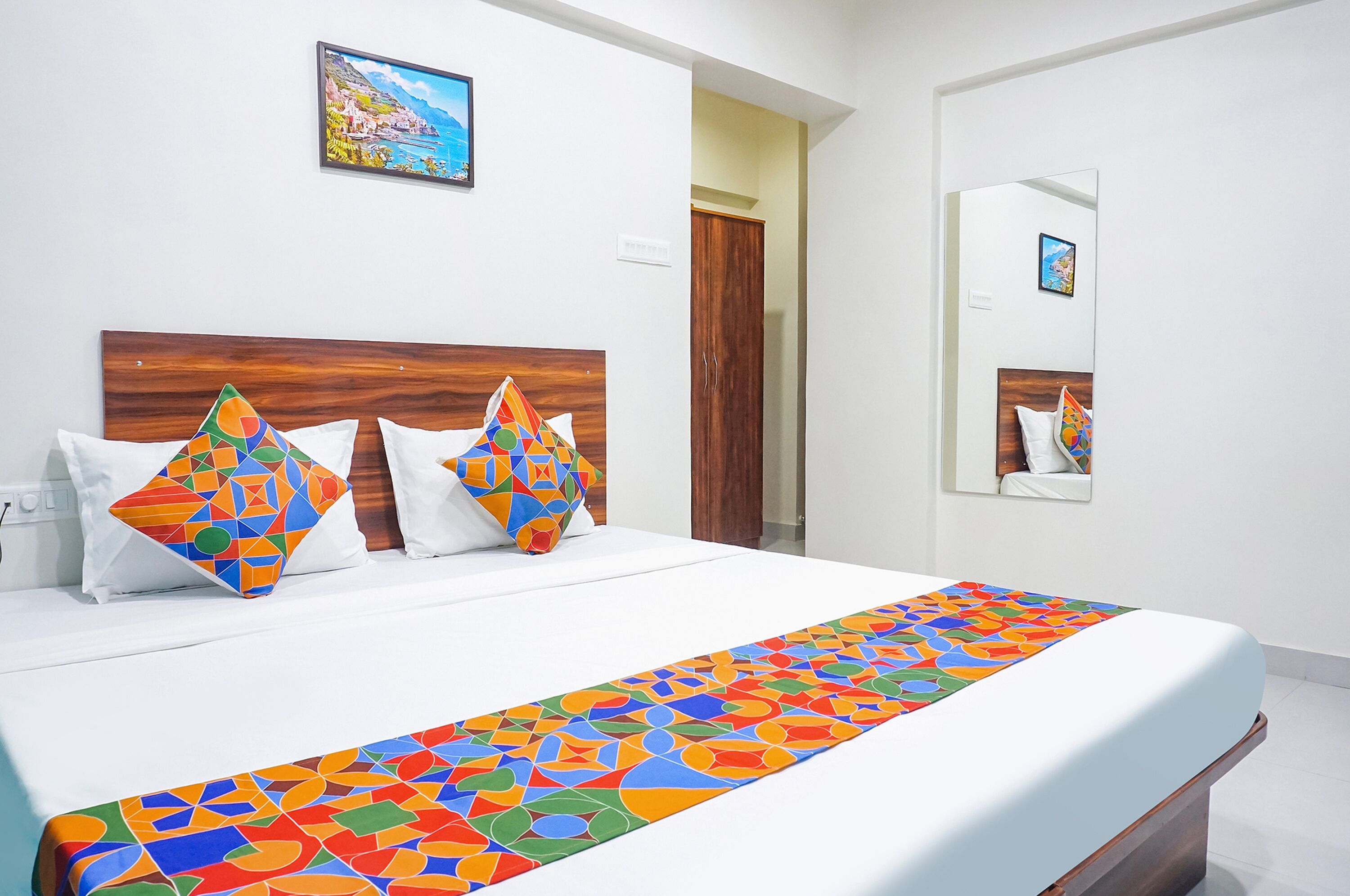 A hotel room with a large bed, colorful geometric pillows, a wooden headboard, a painting on the wall, and a mirror reflecting the room.