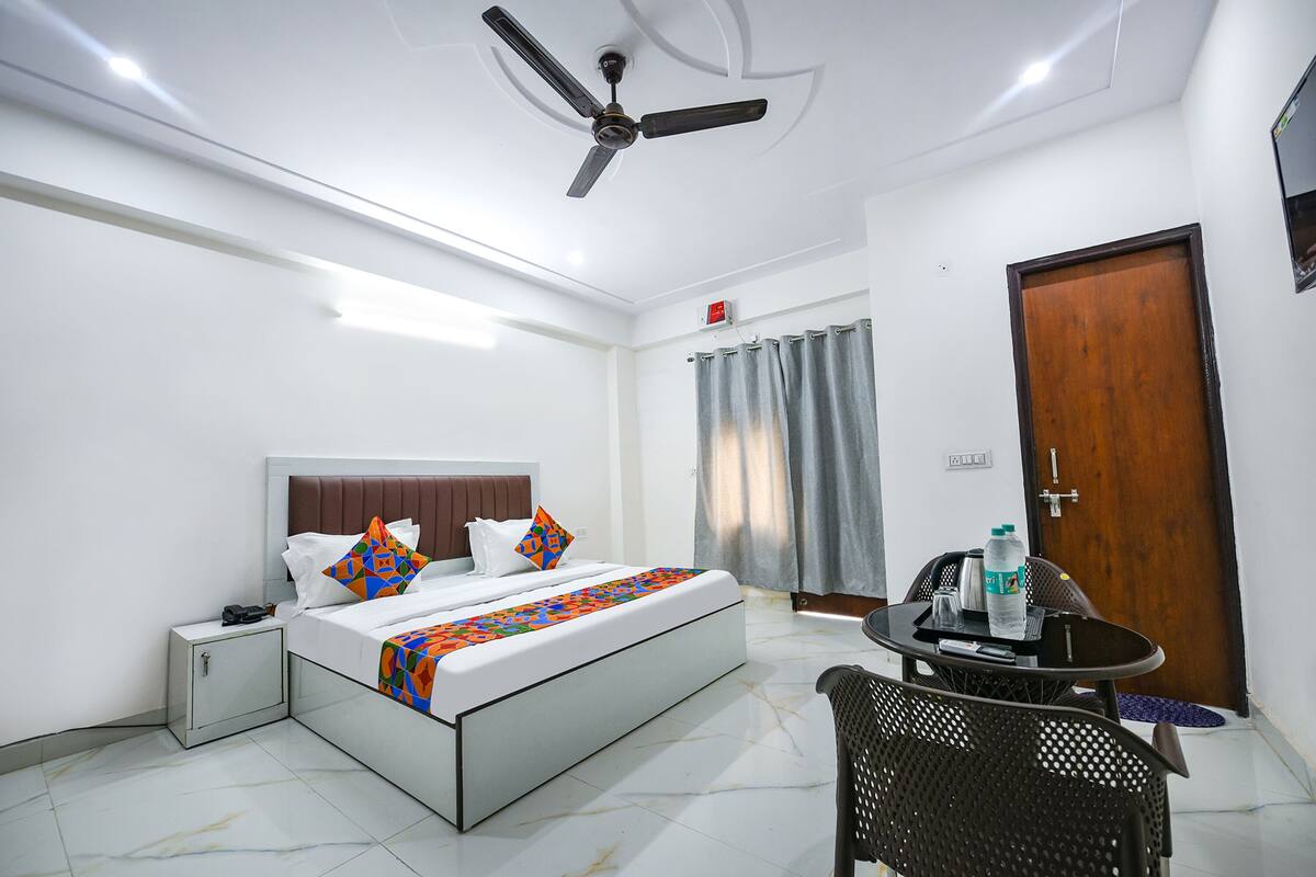A hotel room with a bed, bedside table, chair, and a small table with a bottle and a cup.