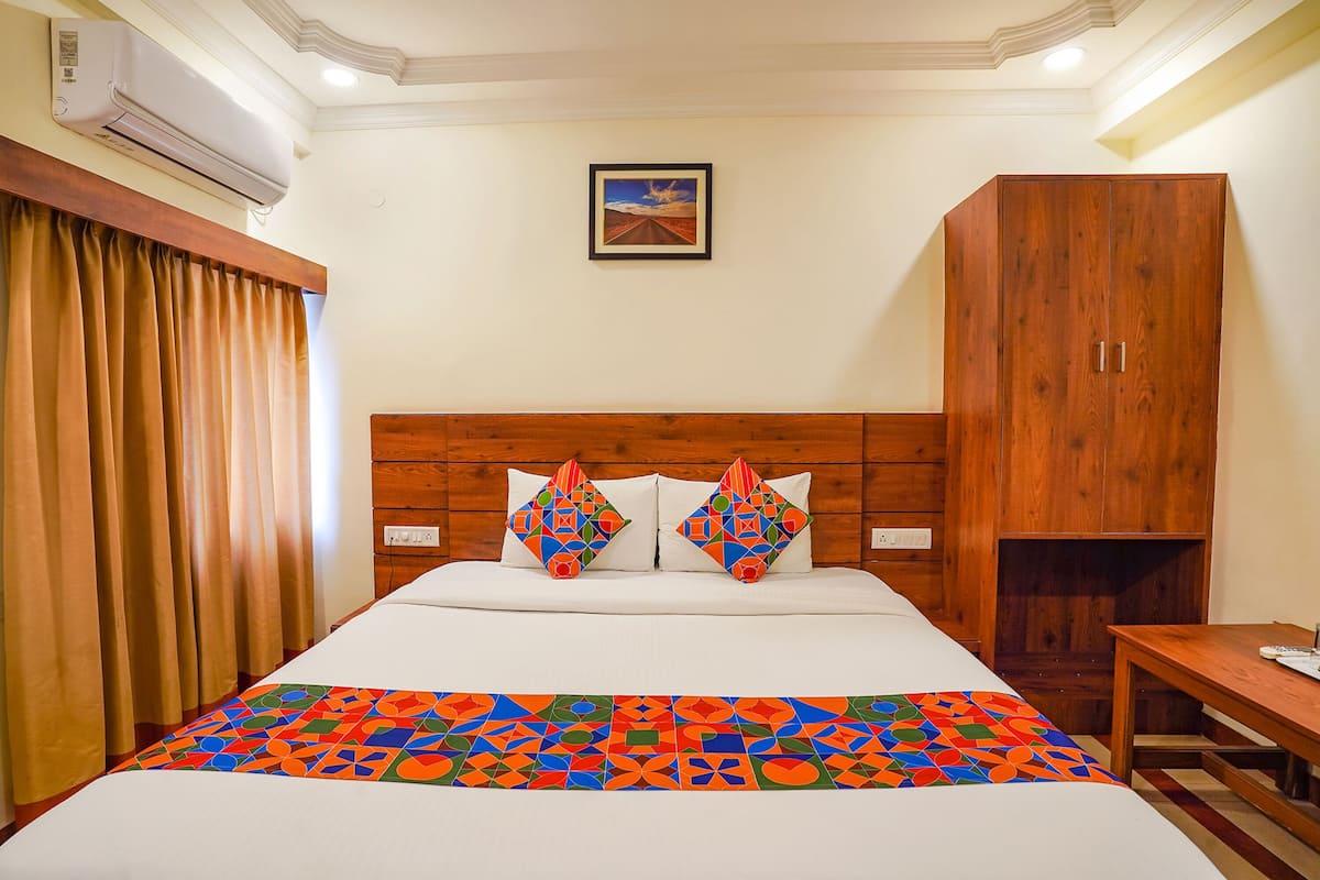 A hotel room with a wooden headboard, a bed with a colorful geometric-patterned bedspread, two decorative pillows, a wooden wardrobe, and a small table with a chair.