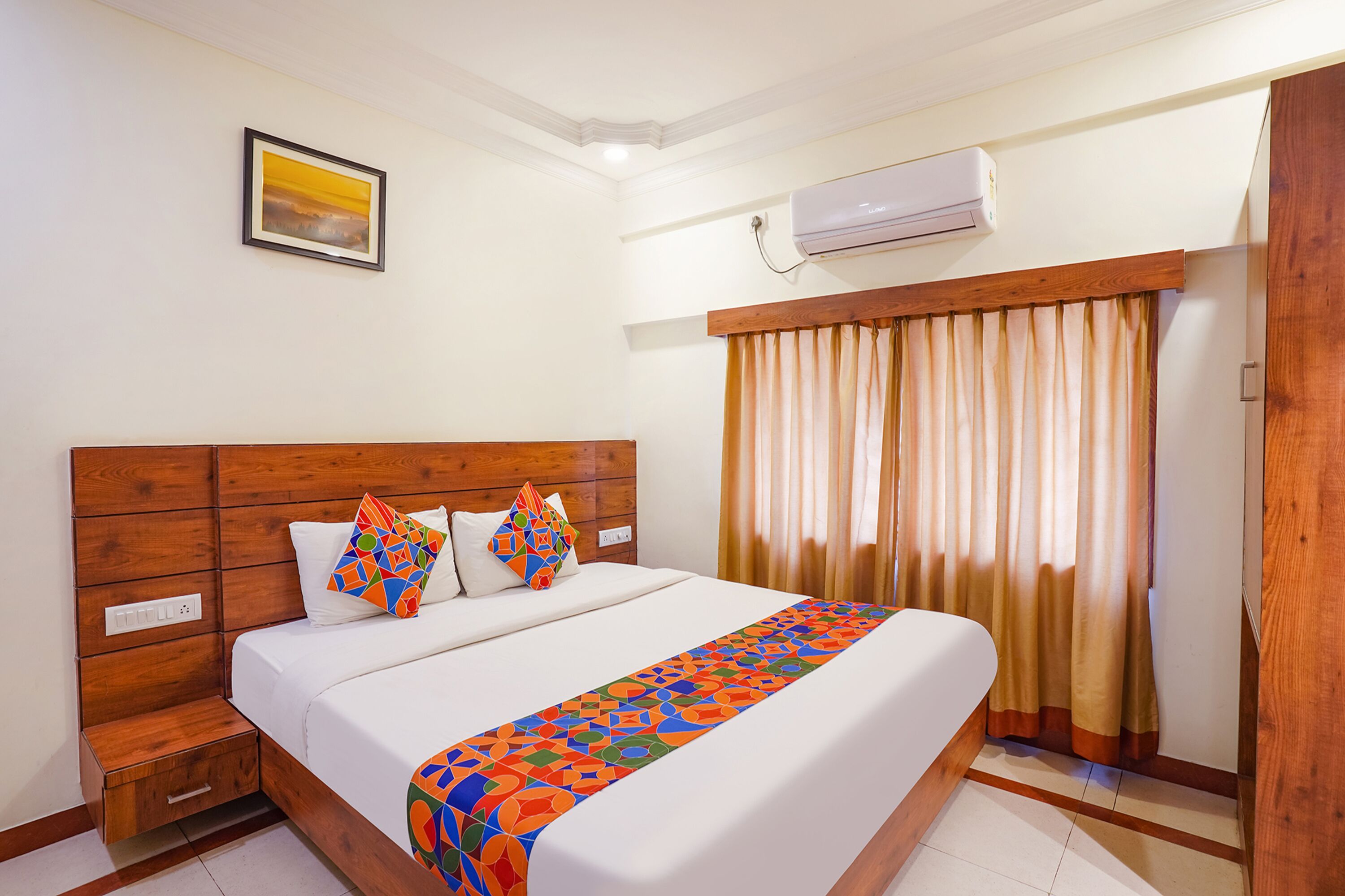 A hotel room with a wooden headboard, a bed with white linens, a colorful throw blanket, a small bedside table, a nightstand, a wall-mounted air conditioner, a framed picture, and a window with curtains.