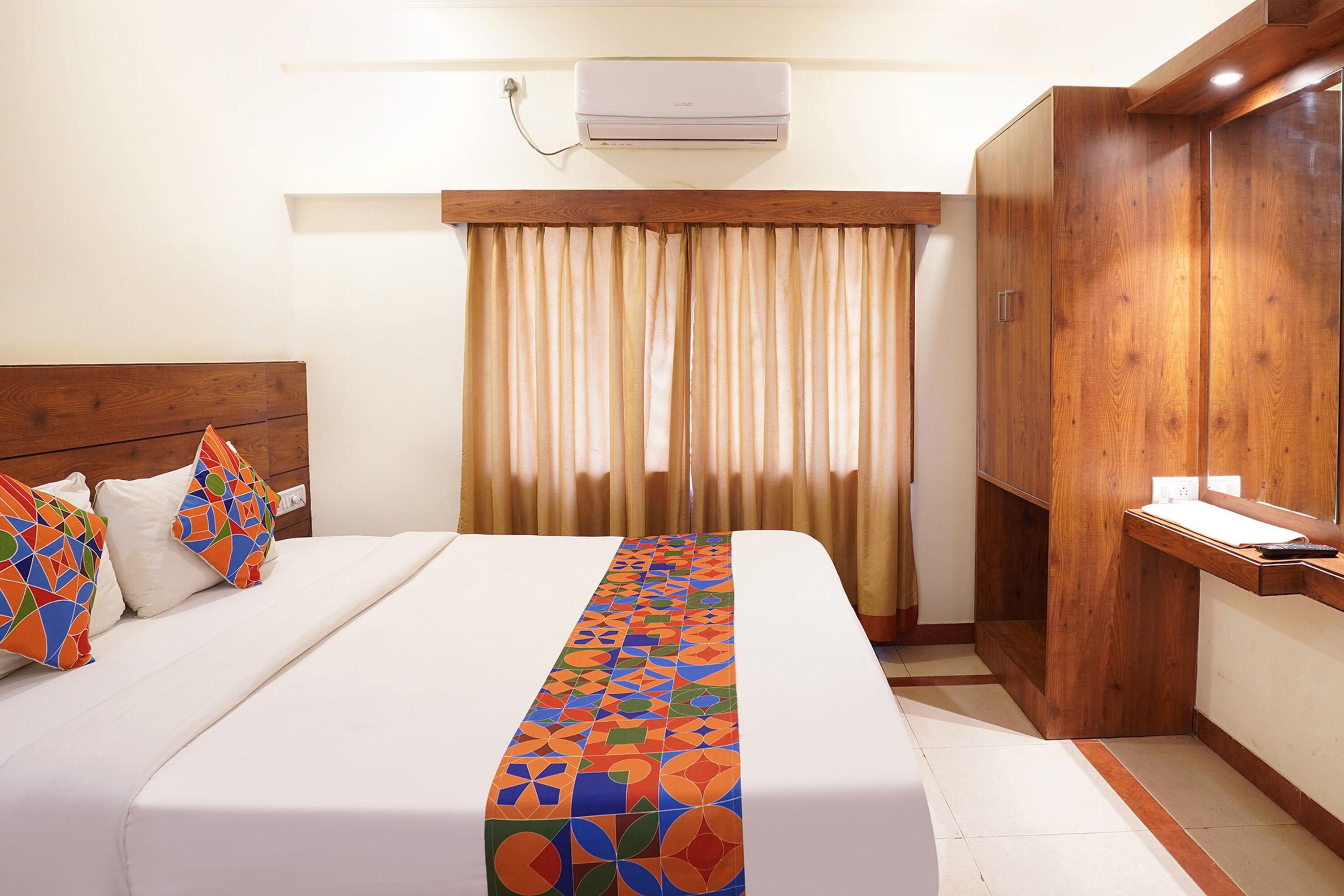 A hotel room with a bed, a wooden wardrobe, a desk, and an air conditioning unit.