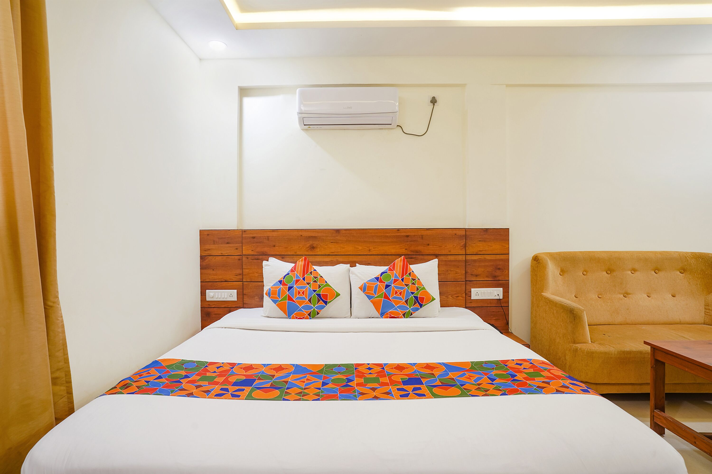 A hotel room with a bed, two decorative pillows, a wooden headboard, a sofa, and an air conditioning unit.