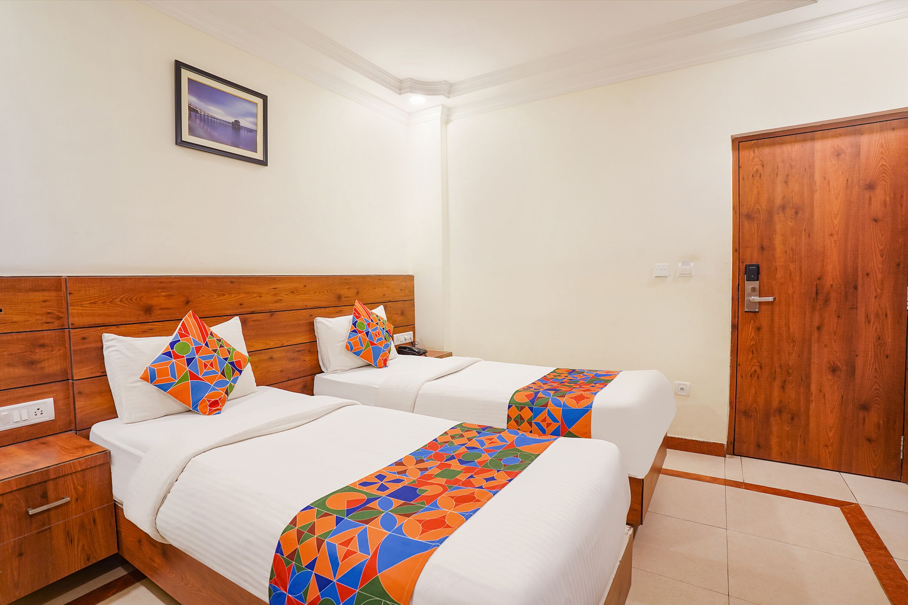 A hotel room with two beds, wooden headboards, white bedding, colorful geometric-patterned bedspreads, a wooden door, and a framed picture on the wall.