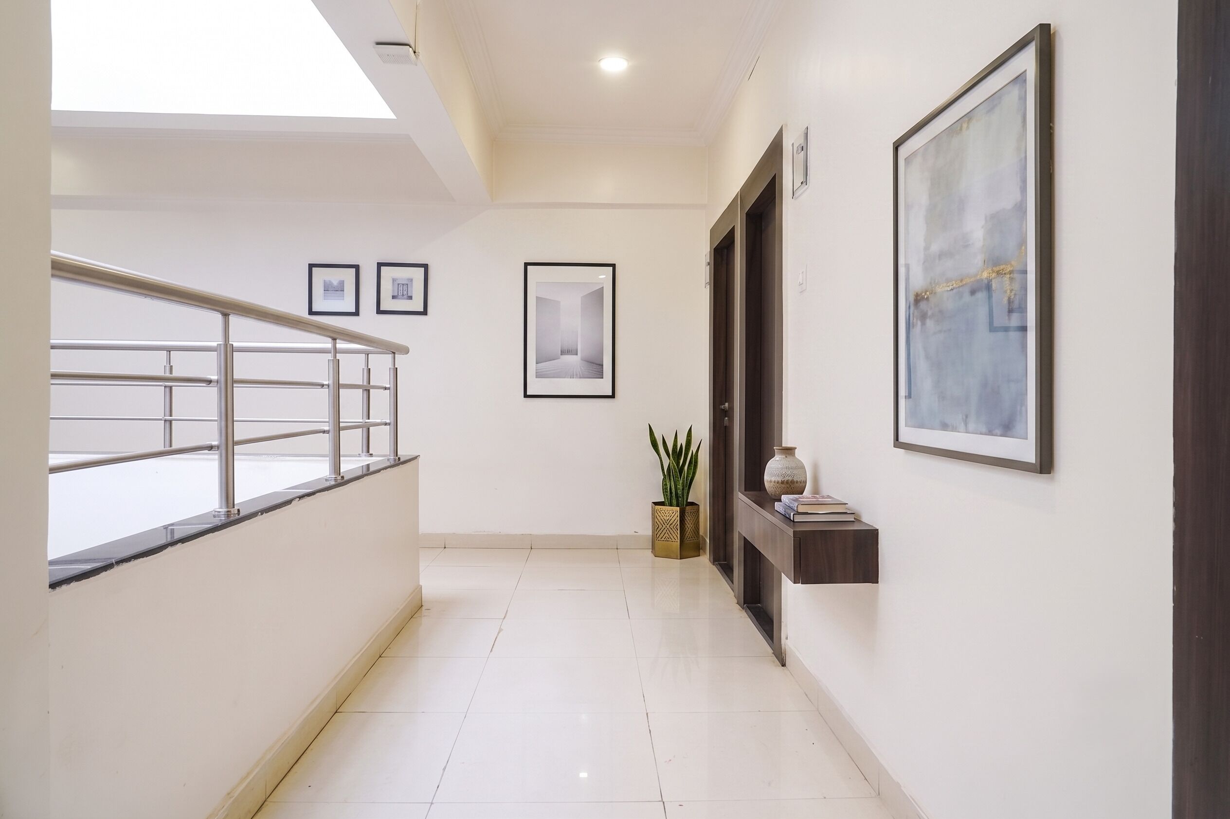 A corridor with a staircase, framed pictures on the walls, a potted plant, and a small shelf with books.