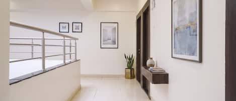 A corridor with a staircase, framed pictures on the walls, a potted plant, and a small shelf with books.