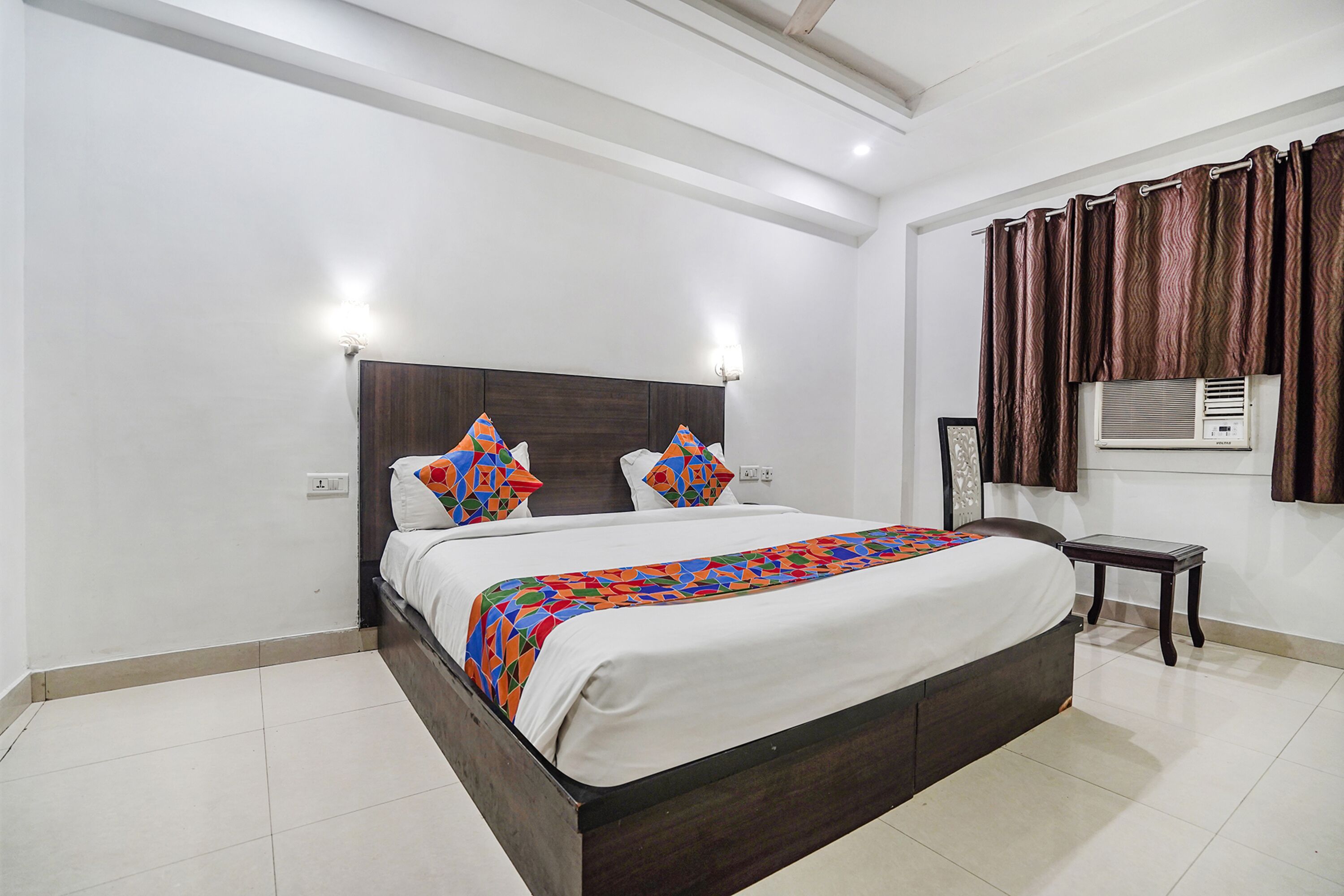 A neatly arranged hotel room with a large bed, a bedside table, a wall-mounted air conditioning unit, and a curtain.