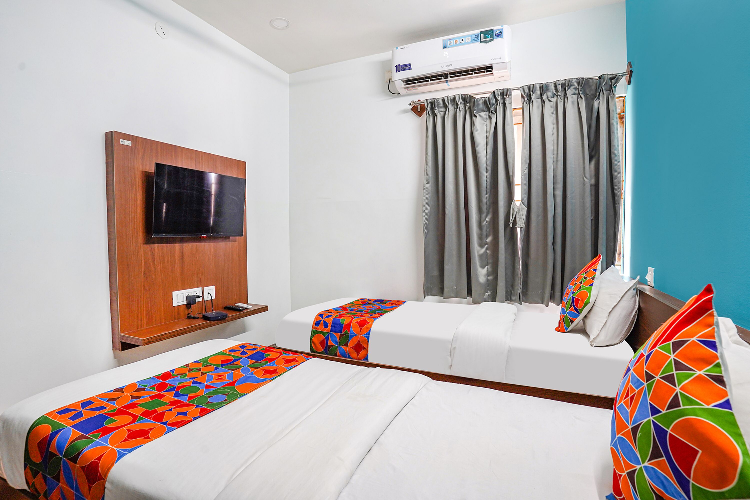 A hotel room with two beds, a TV mounted on the wall, an air conditioning unit, and a window with curtains.