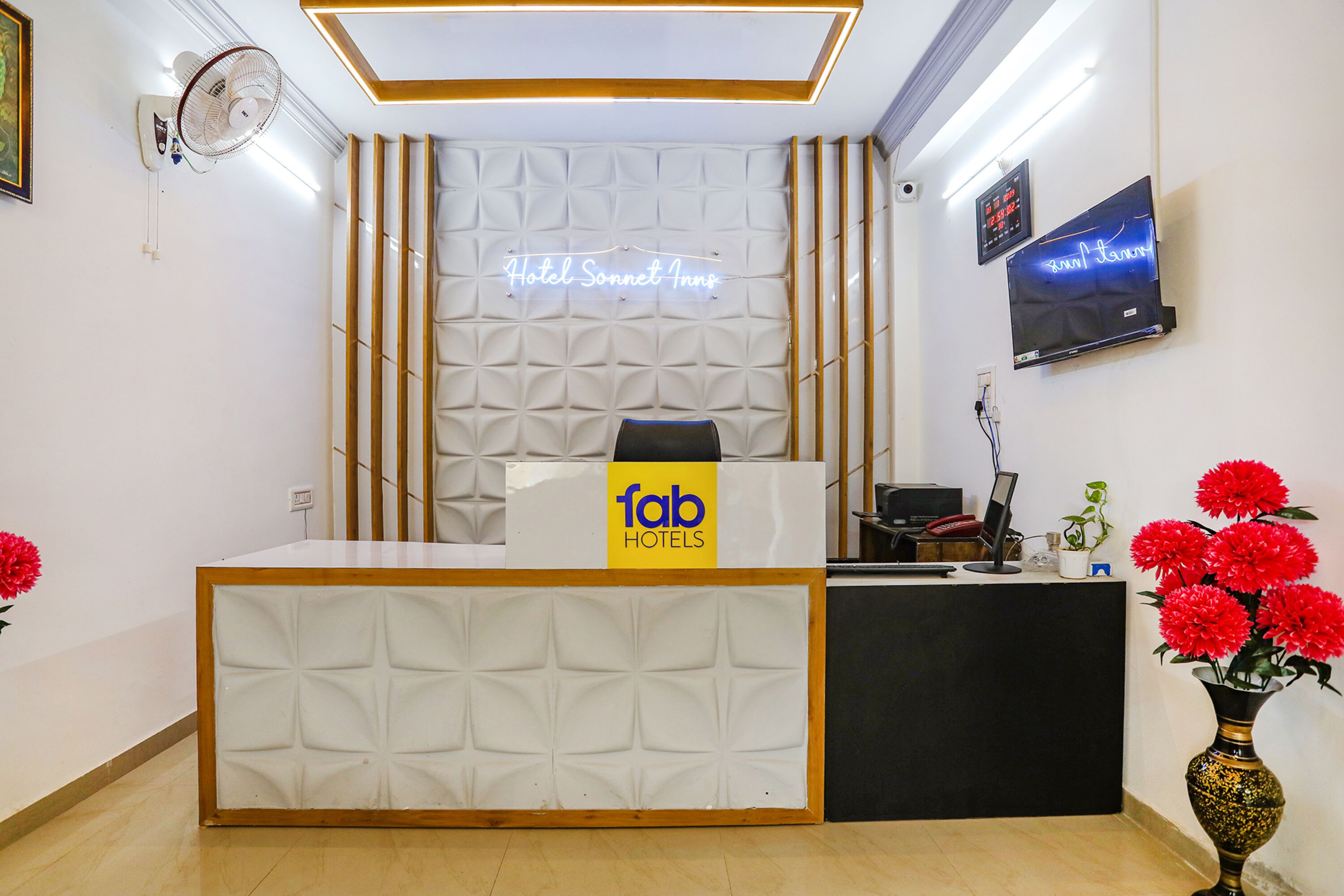 Una recepción de hotel con un letrero de 'fab hotels', un letrero de neón sobre el mostrador, un televisor de pantalla plana y un jarrón con flores rojas.