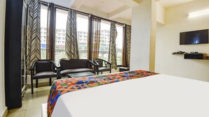 Una habitación de hotel con cama, dos sillas, televisor y ventanas amplias con cortinas.