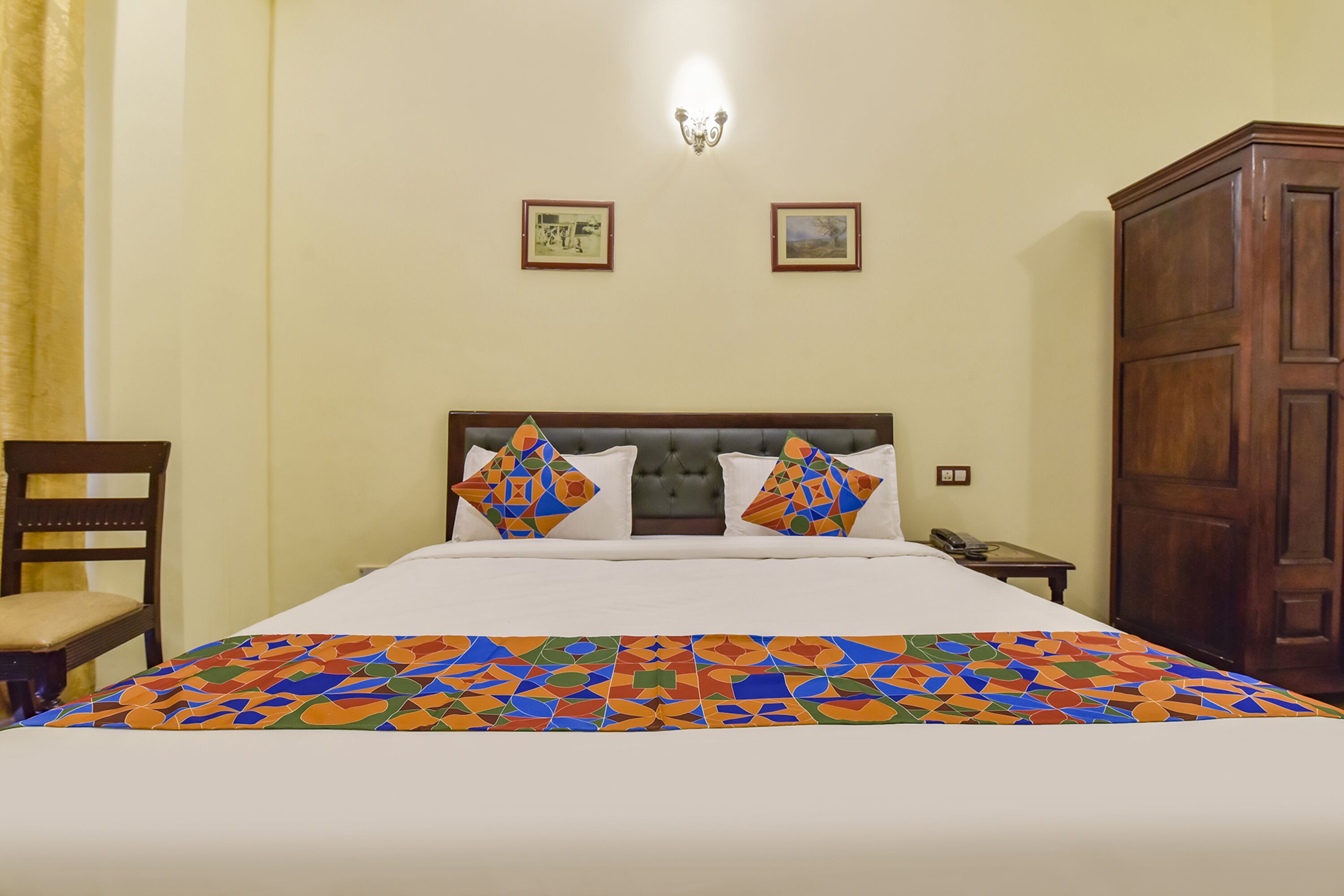 A neatly made bed with a colorful geometric-patterned bedspread, a wooden nightstand with a telephone, and a wooden wardrobe.