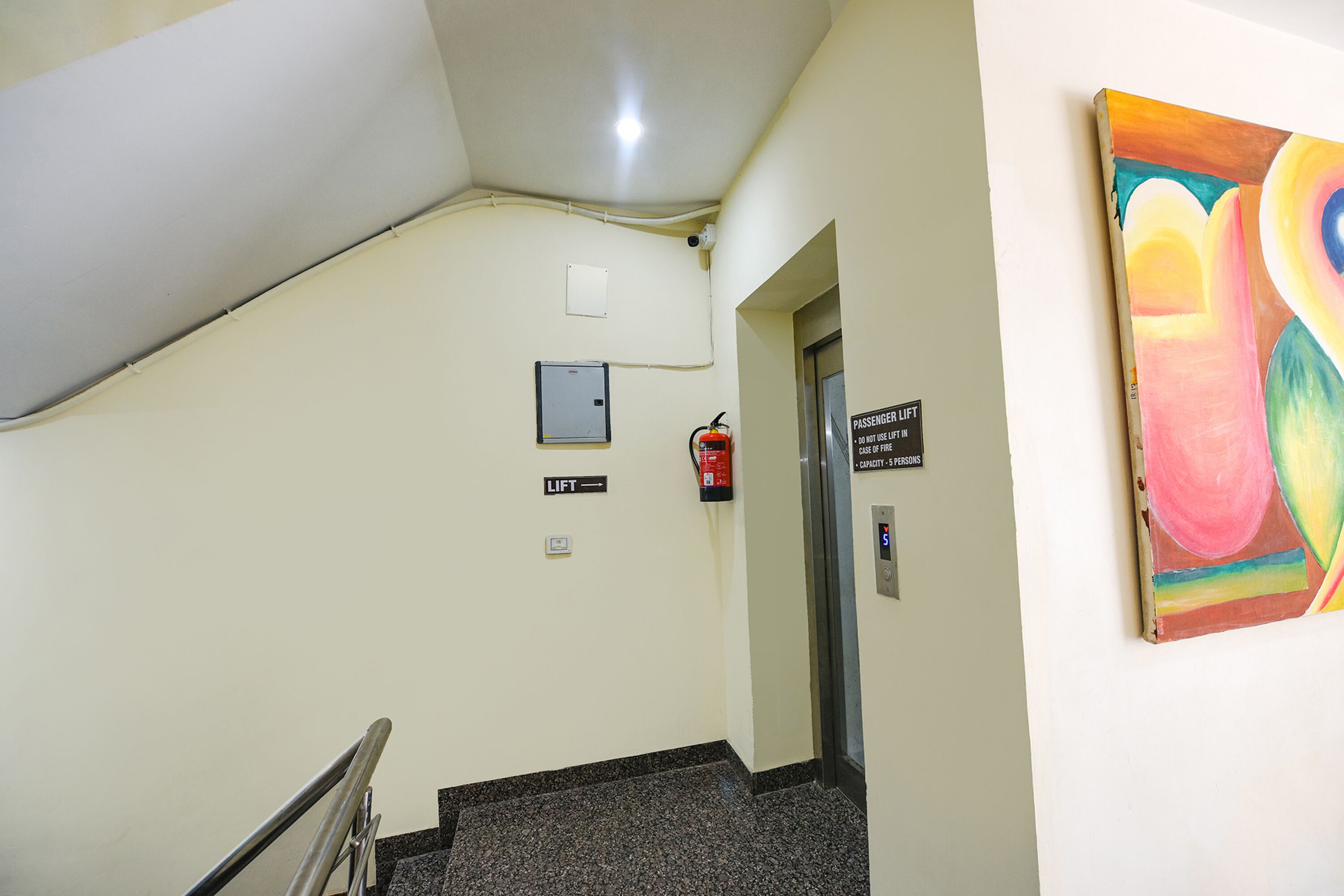 Un escalier avec un extincteur, un panneau indiquant la présence d’un ascenseur et un tableau accroché au mur.