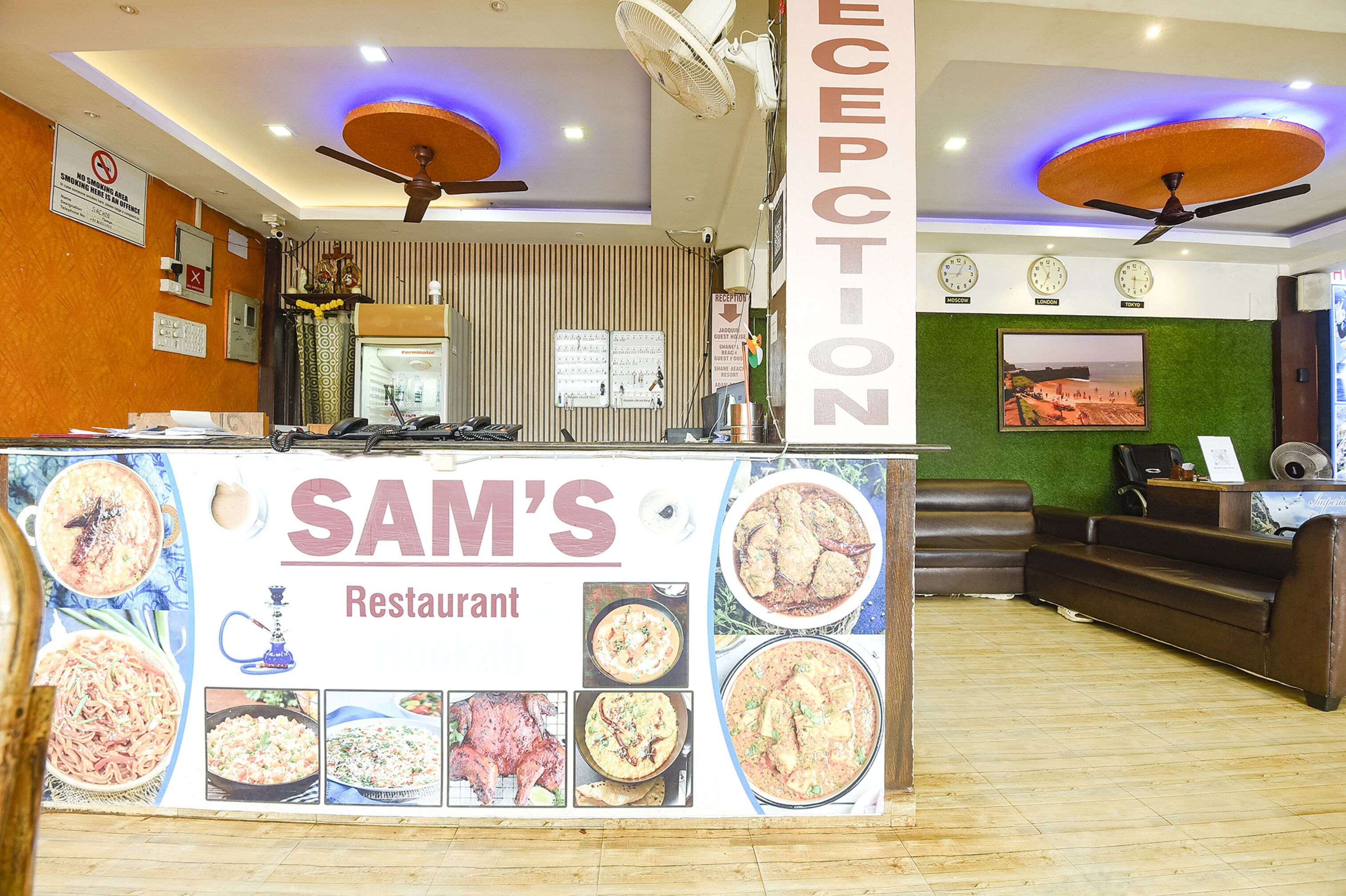 A restaurant reception area with a counter displaying food images, ceiling fans, clocks, and a green wall with a picture.