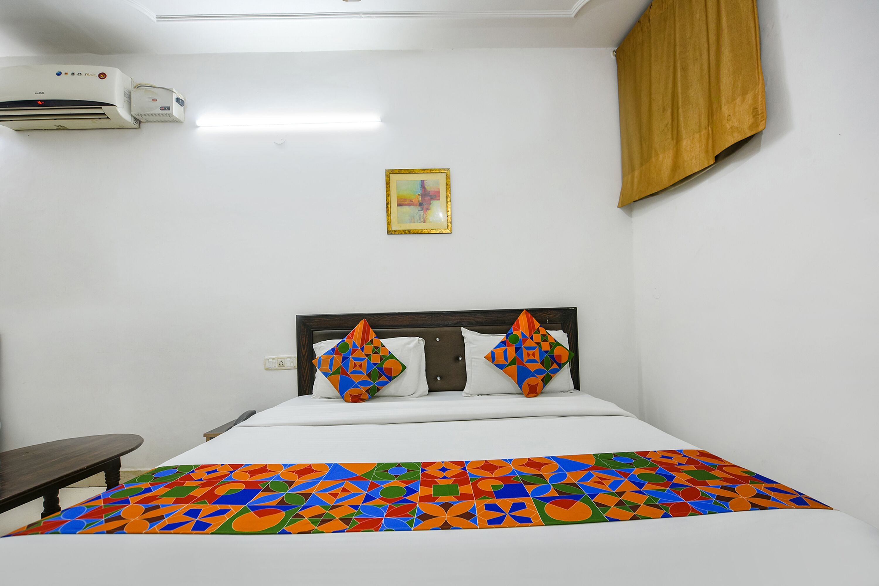A hotel room with a bed, a nightstand, a framed picture on the wall, and an air conditioning unit.