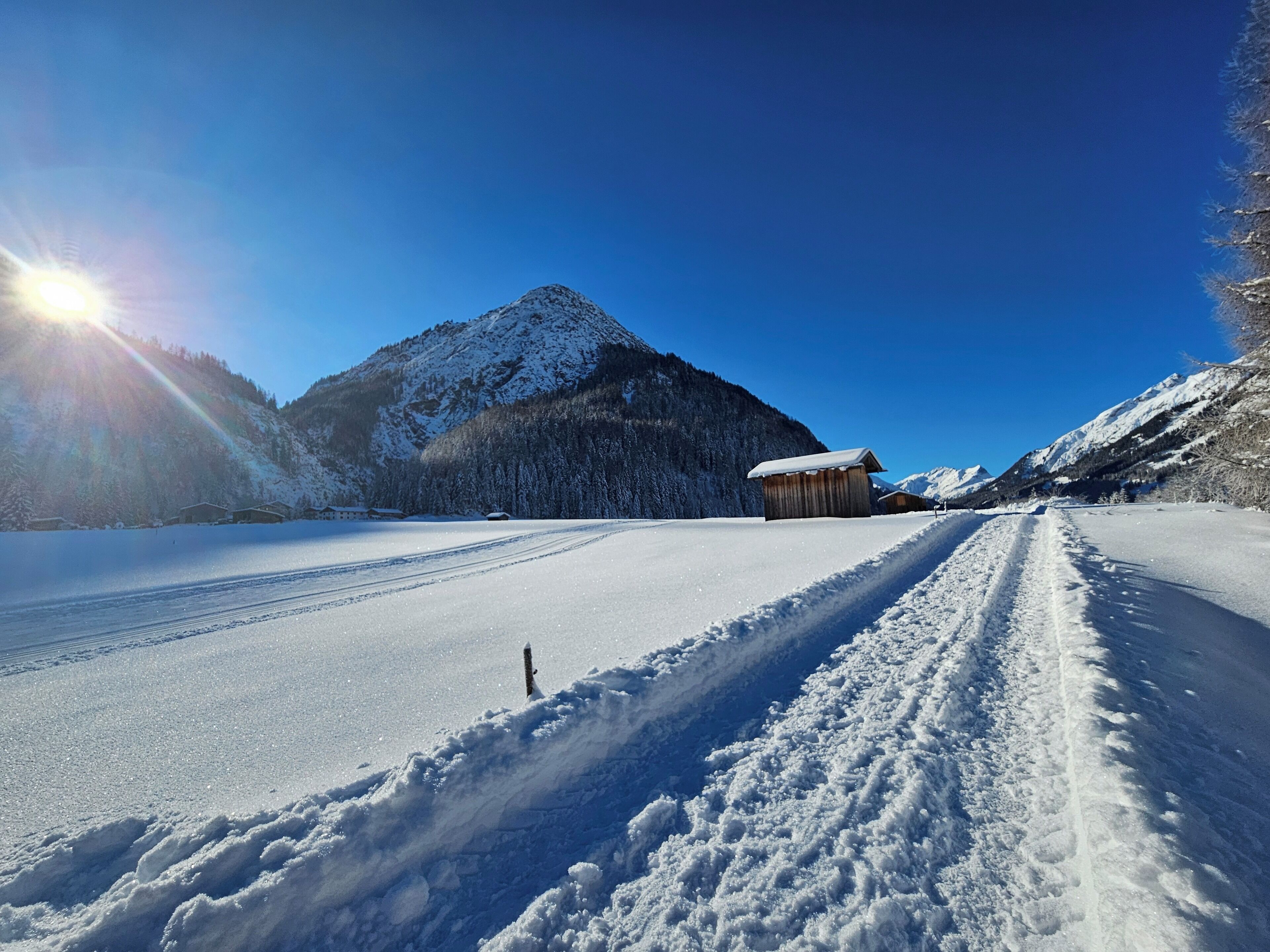 A snow-covered road with a wooden cabin and a mountain in the background.