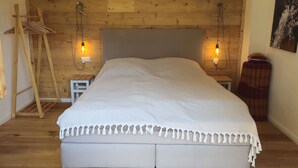 A bedroom with a wooden ceiling, a bed with a white bedspread, two bedside lamps, and a wooden bench.