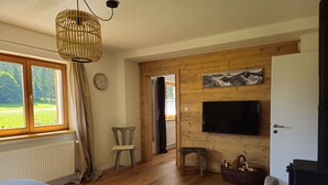 A cozy living room with a wooden floor, a round table with a glass, a TV mounted on the wall, and a window with curtains.