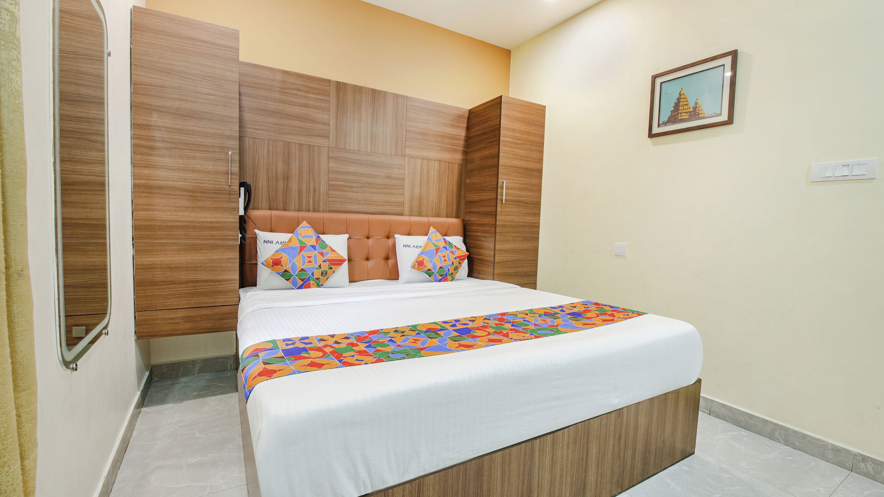 Une chambre d’hôtel avec un lit, une tête de lit en bois, deux oreillers, une couvre-lit colorée et un tableau encadré au mur.
