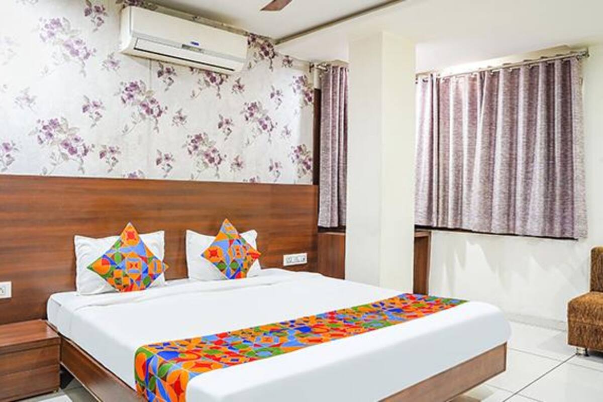 A hotel room with a wooden headboard, floral wallpaper, a bed with colorful bedding, and an air conditioning unit.