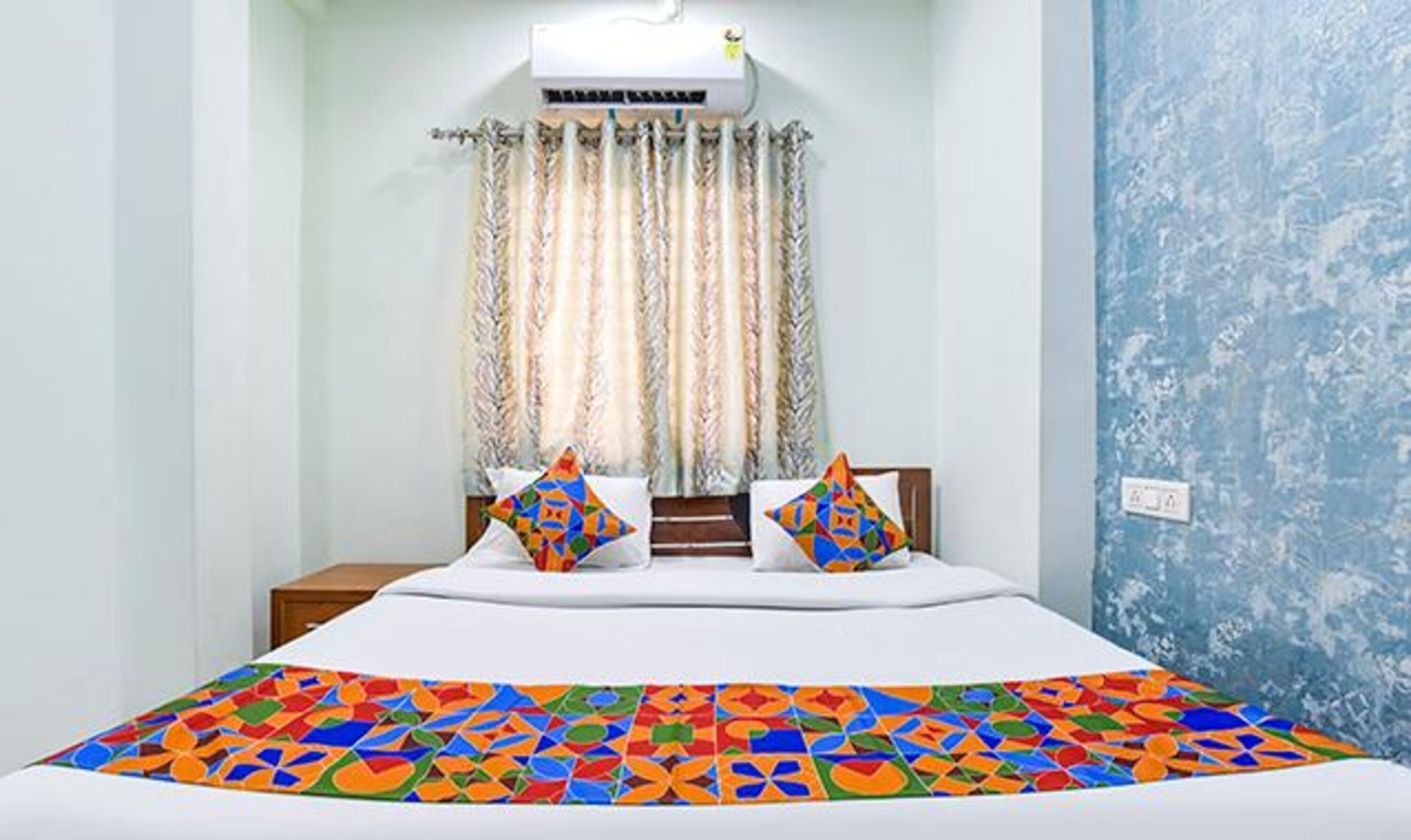 A bedroom with a bed, two pillows, a window with curtains, an air conditioning unit, and a wall with a textured pattern.