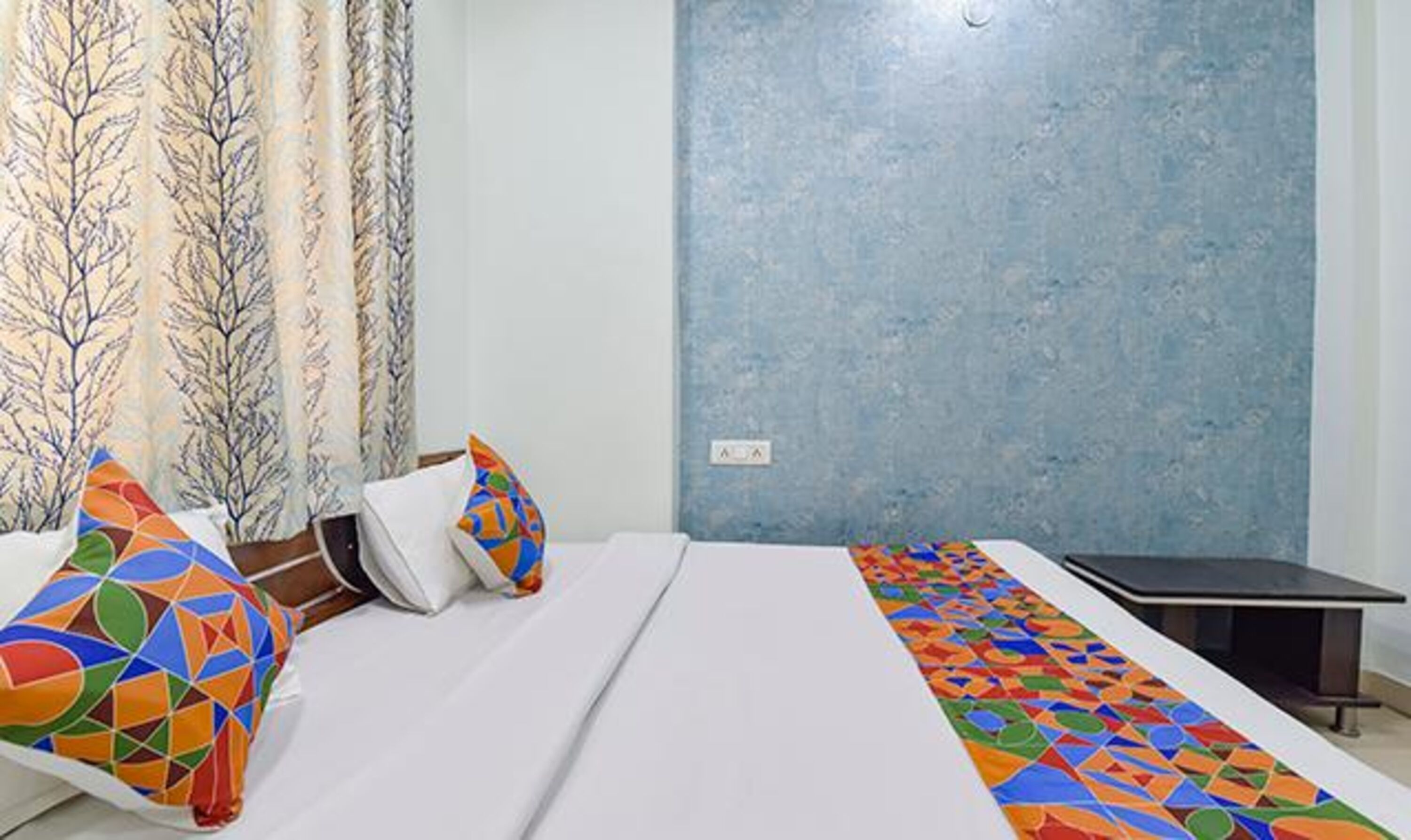 A hotel room with a bed, pillows, a desk, and a patterned curtain.