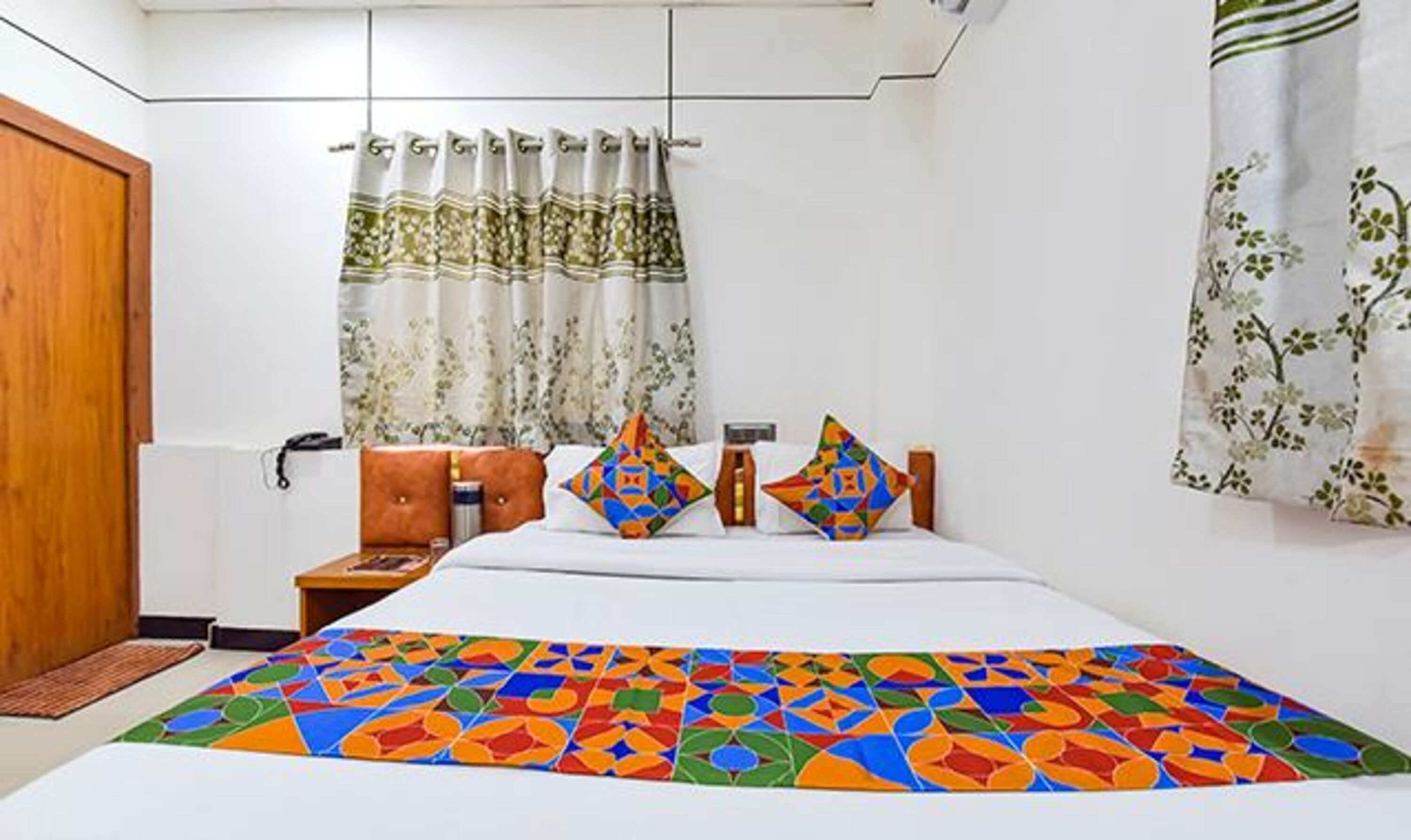 A hotel room with a bed, pillows, a nightstand, a wooden door, and a window with curtains.