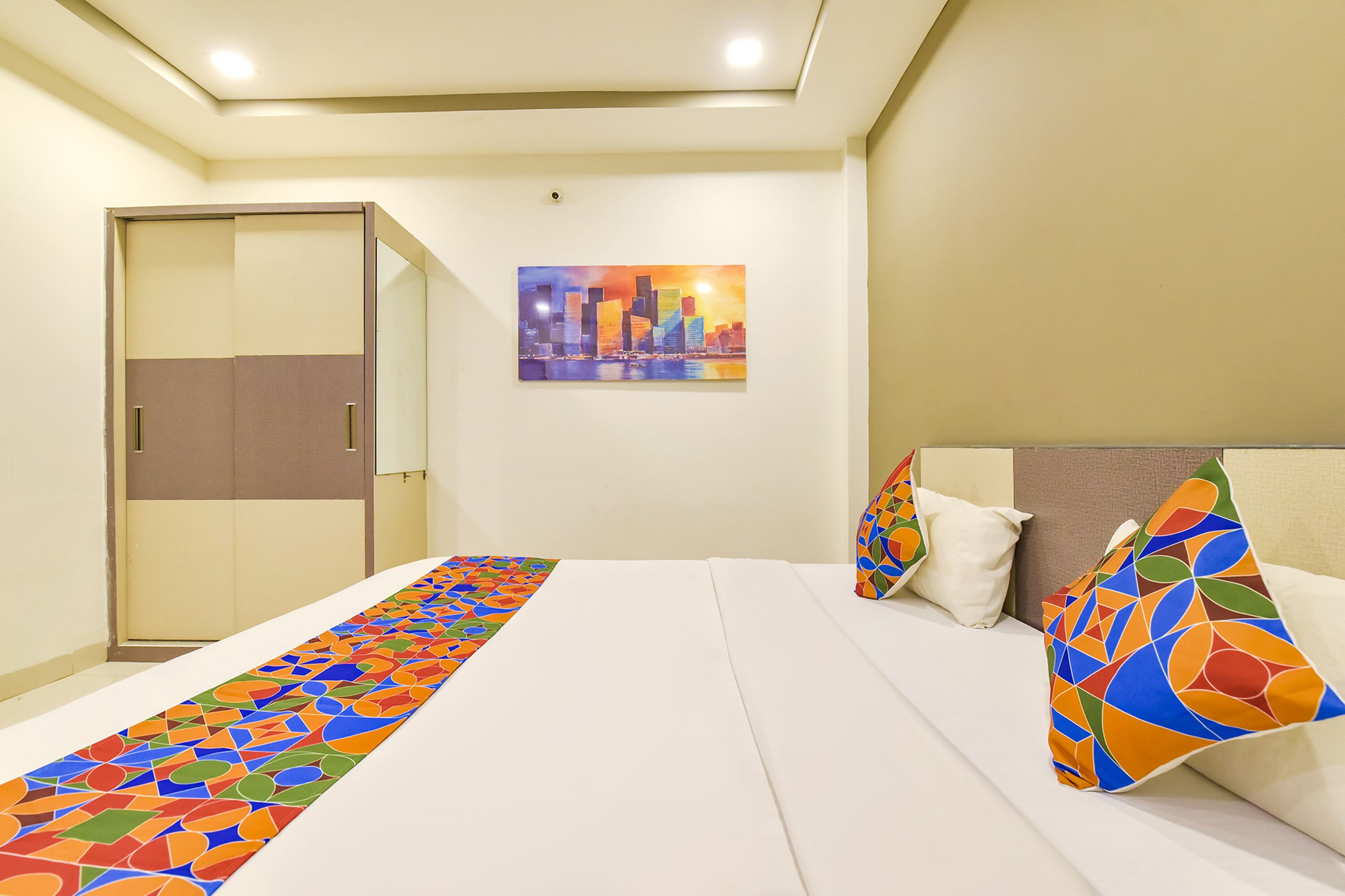 A hotel room with a bed, a colorful runner, a cityscape painting, and a wardrobe.