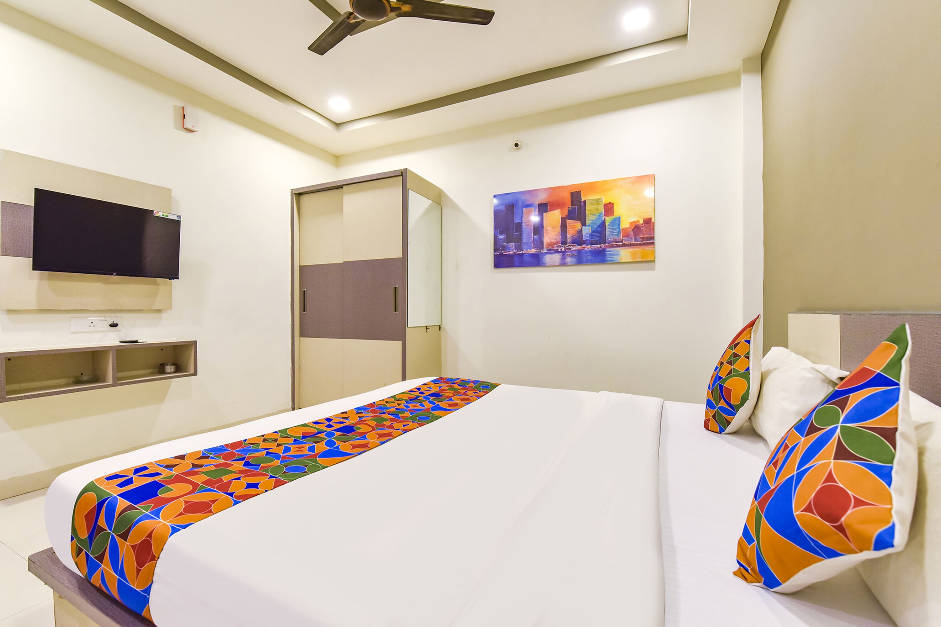 A hotel room with a bed, a television mounted on the wall, a painting, and a ceiling fan.