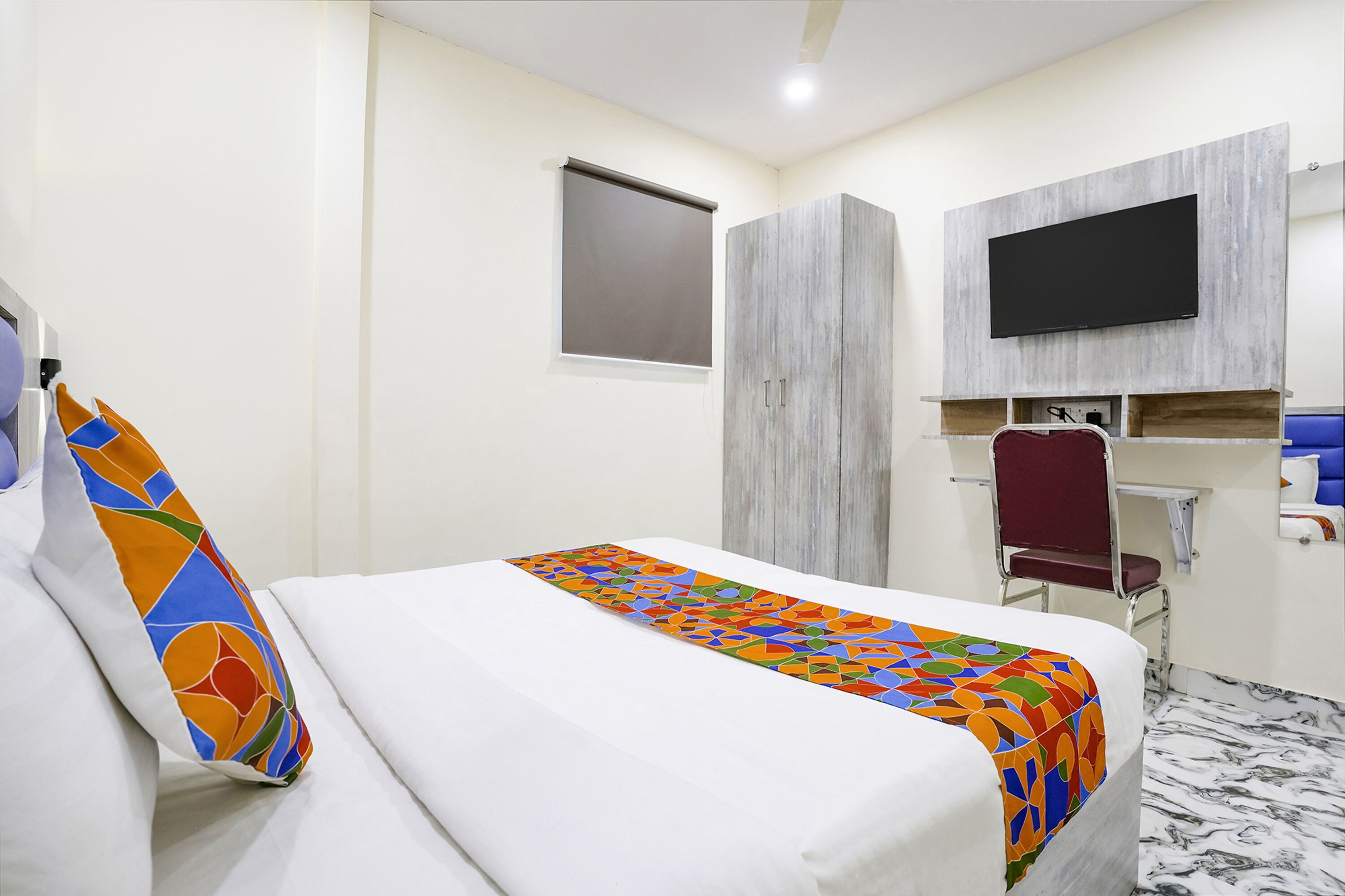 A hotel room with a bed, a TV mounted on the wall, a desk with a chair, and a window with a blind.