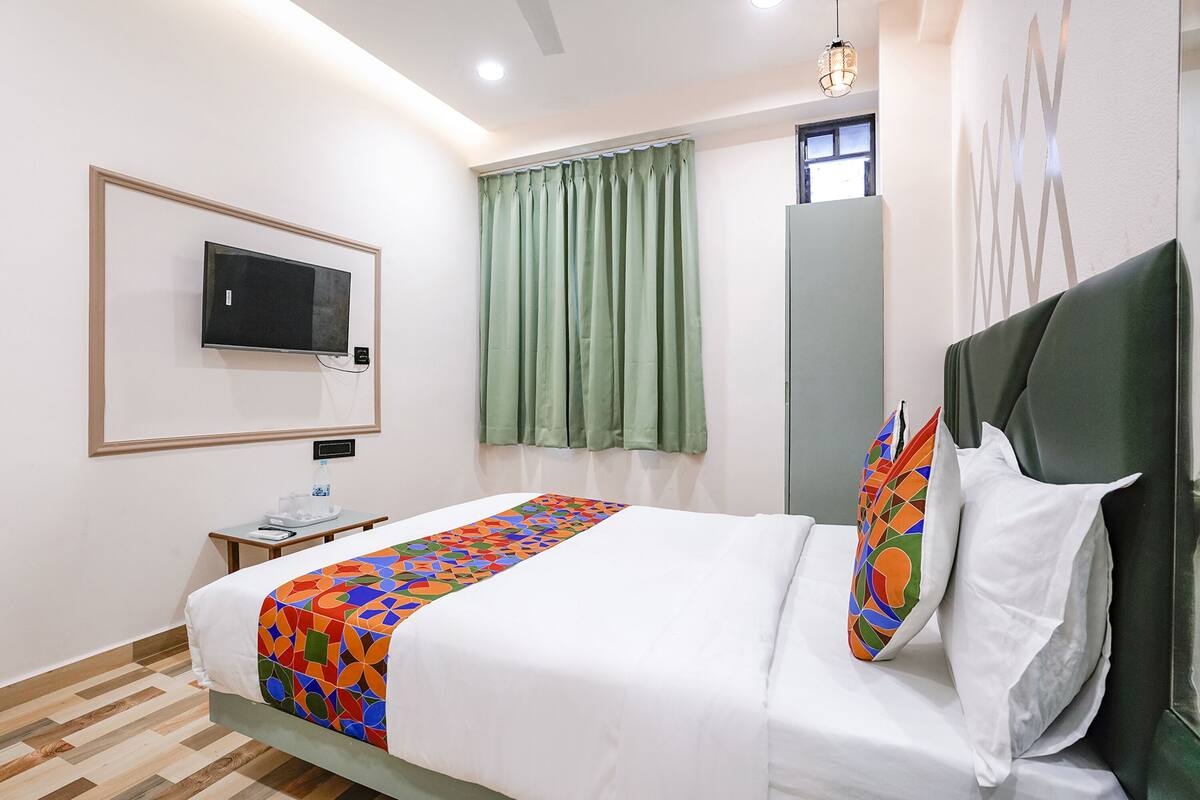 A hotel room with a bed, a TV mounted on the wall, a window with curtains, a bedside table, and a mirror.