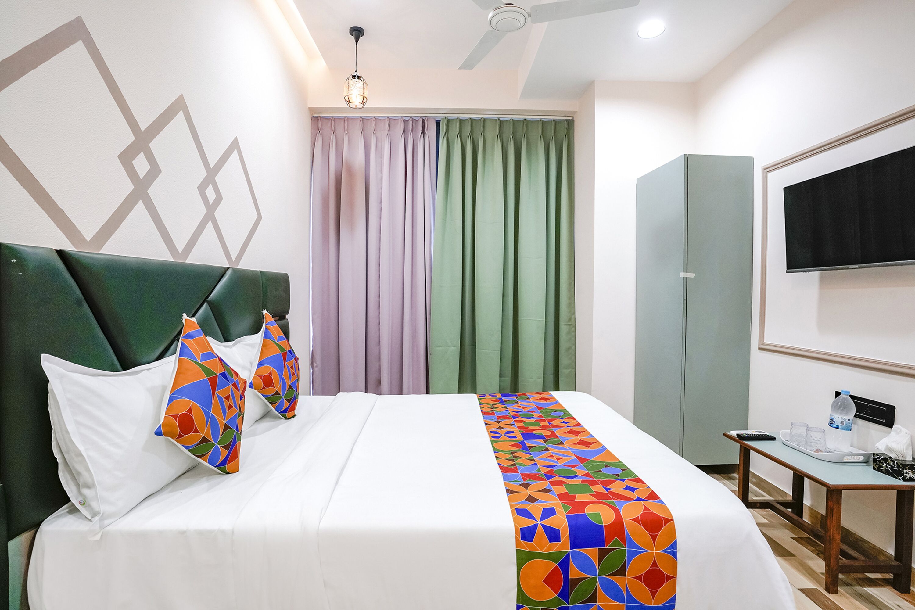 A hotel room with a bed, pillows, a blanket, a TV, a wardrobe, and a table with items on it.