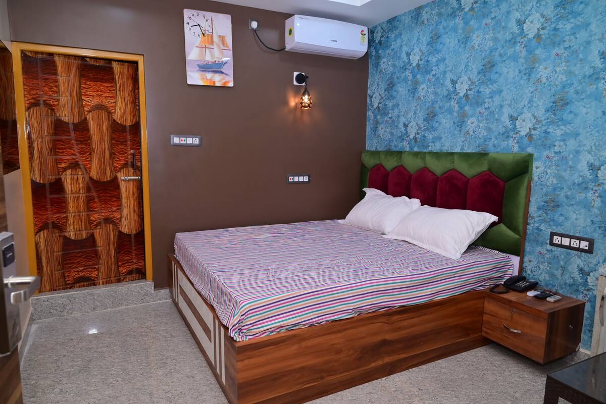 Une chambre à coucher avec un lit en bois, une tête de lit recouverte d’un tissu vert, une couvre-lit à rayures et une table de chevet avec un téléphone et un chargeur.