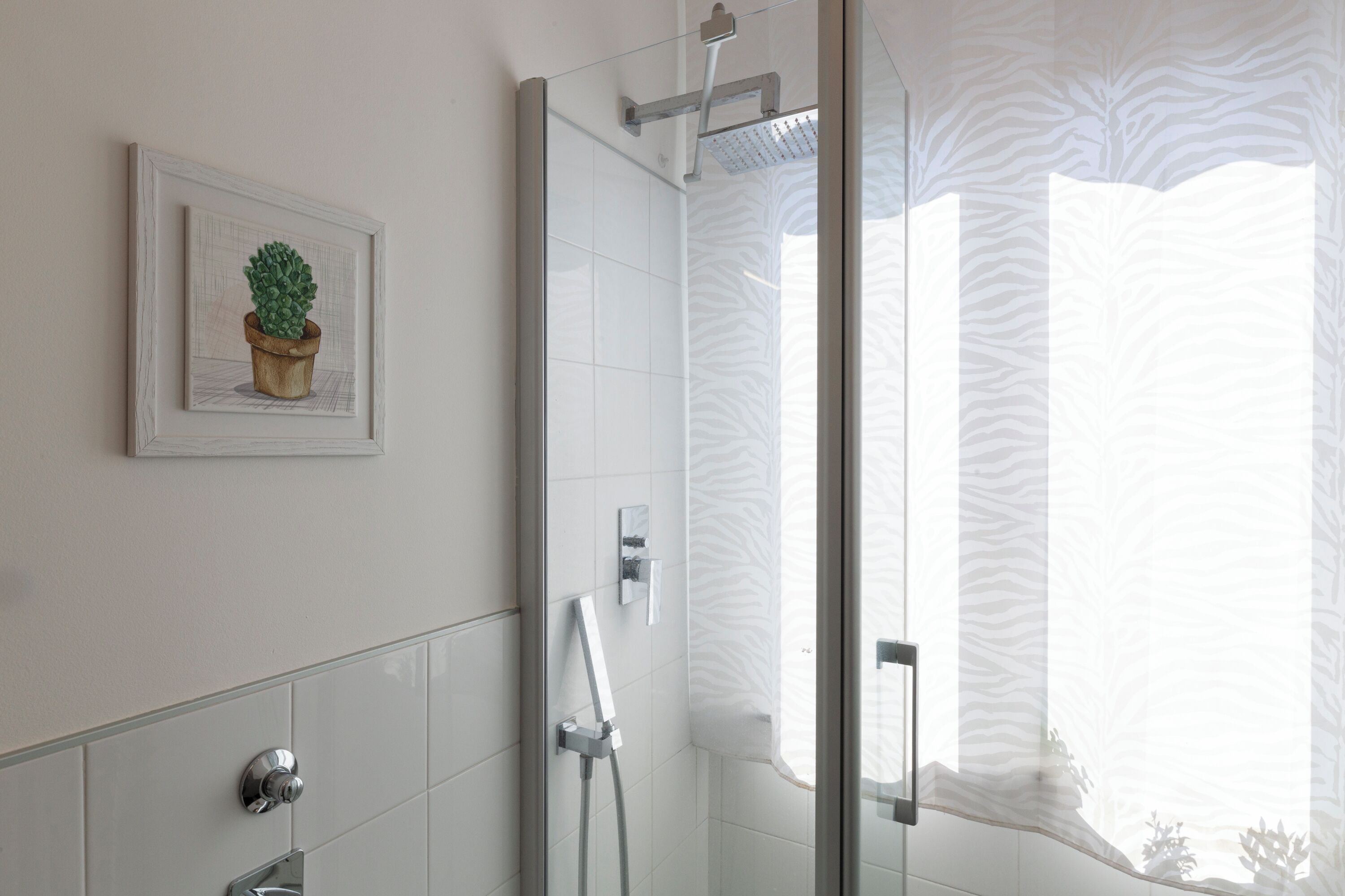 Un bagno con box doccia in vetro, una pianta in vaso sulla parete e una finestra con tenda a motivi zebrati.