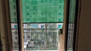 Une vue à travers une fenêtre avec des stores verts, un balcon doté d’une rambarde en métal et un bâtiment en arrière-plan.
