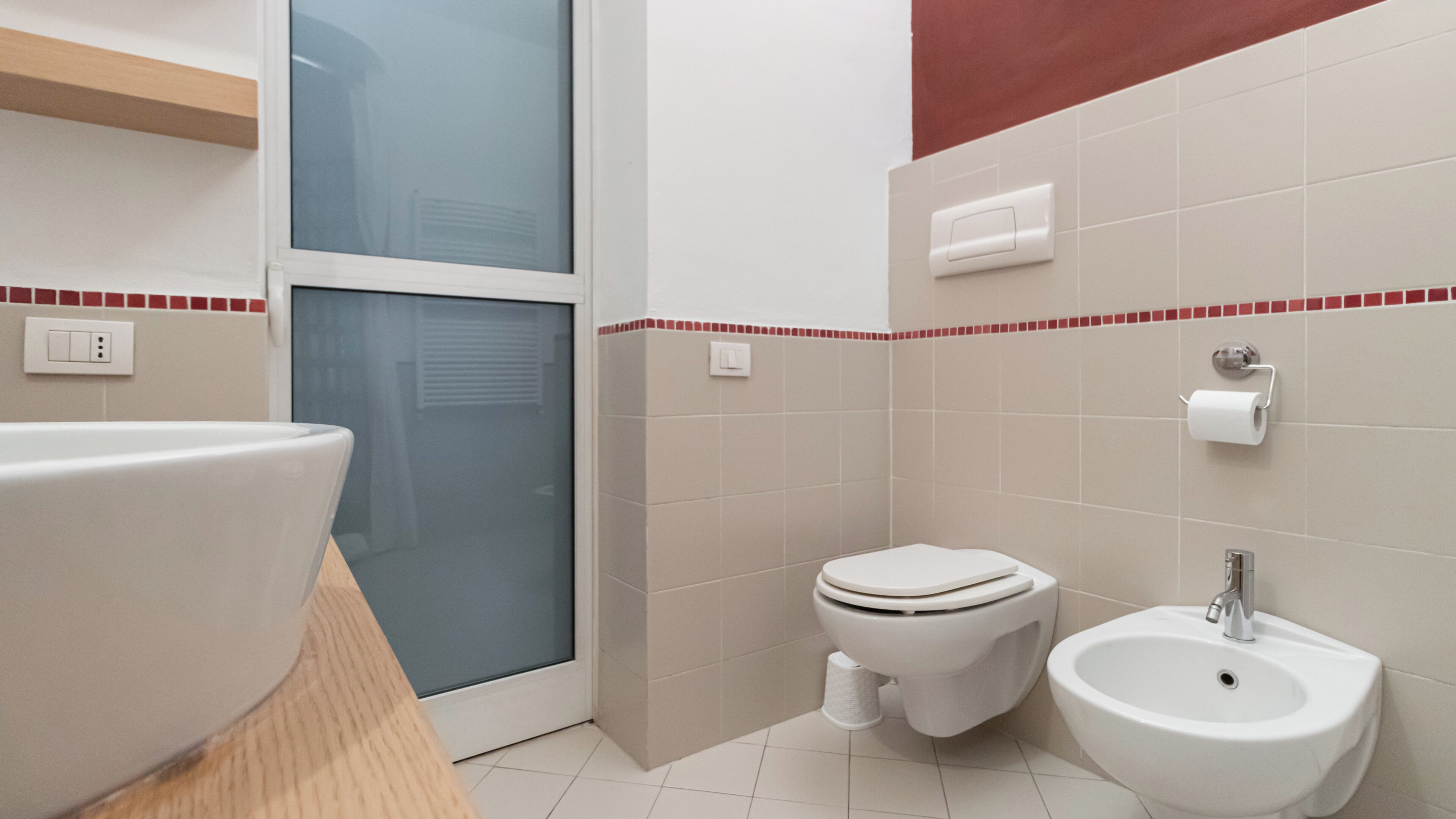 A bathroom with a large white sink, a toilet, and a bidet. There is a window with frosted glass and a red-tiled border.